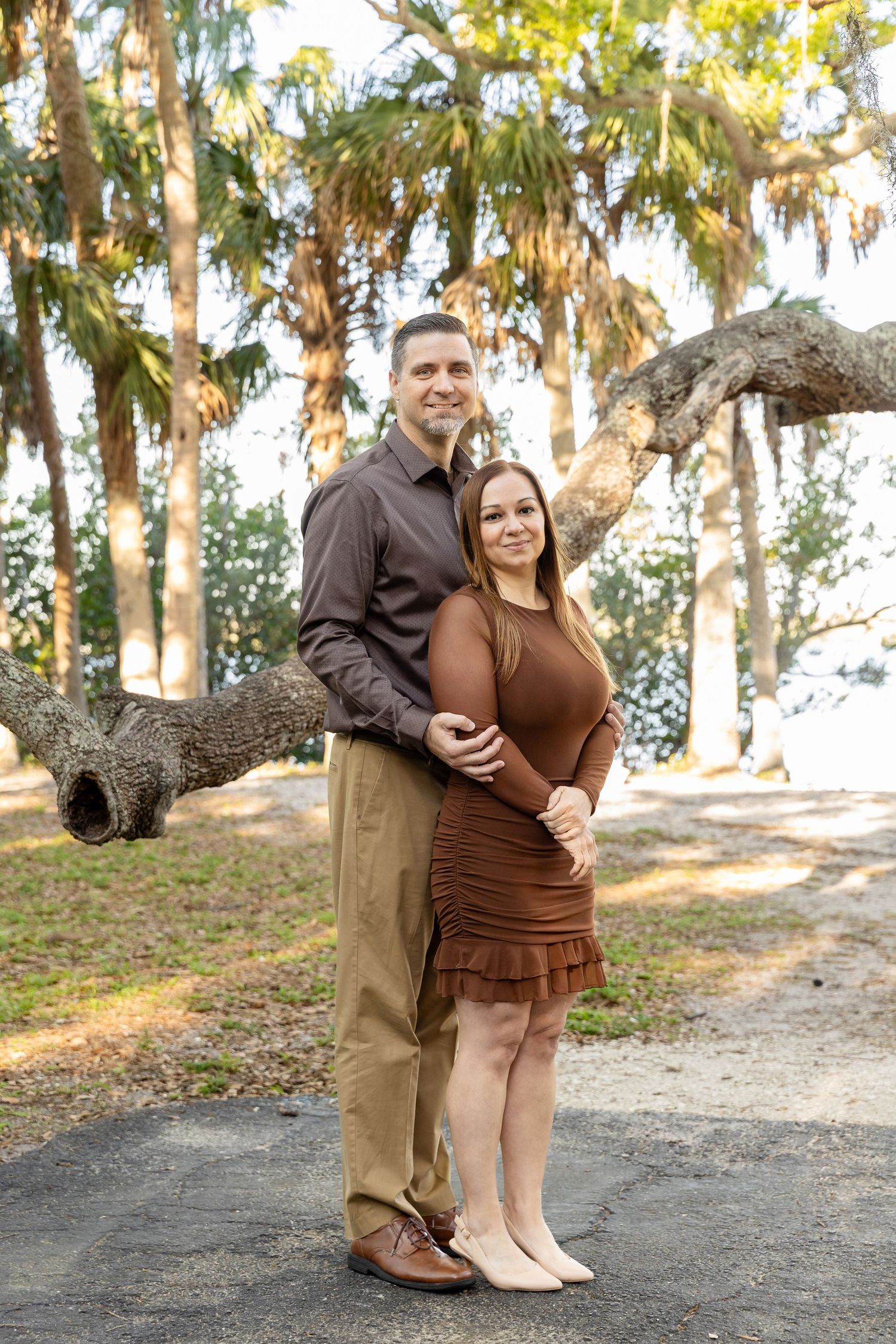 Couple posing outdoors among tall palm trees, wearing coordinating brown outfits, smiling warmly in a park setting.