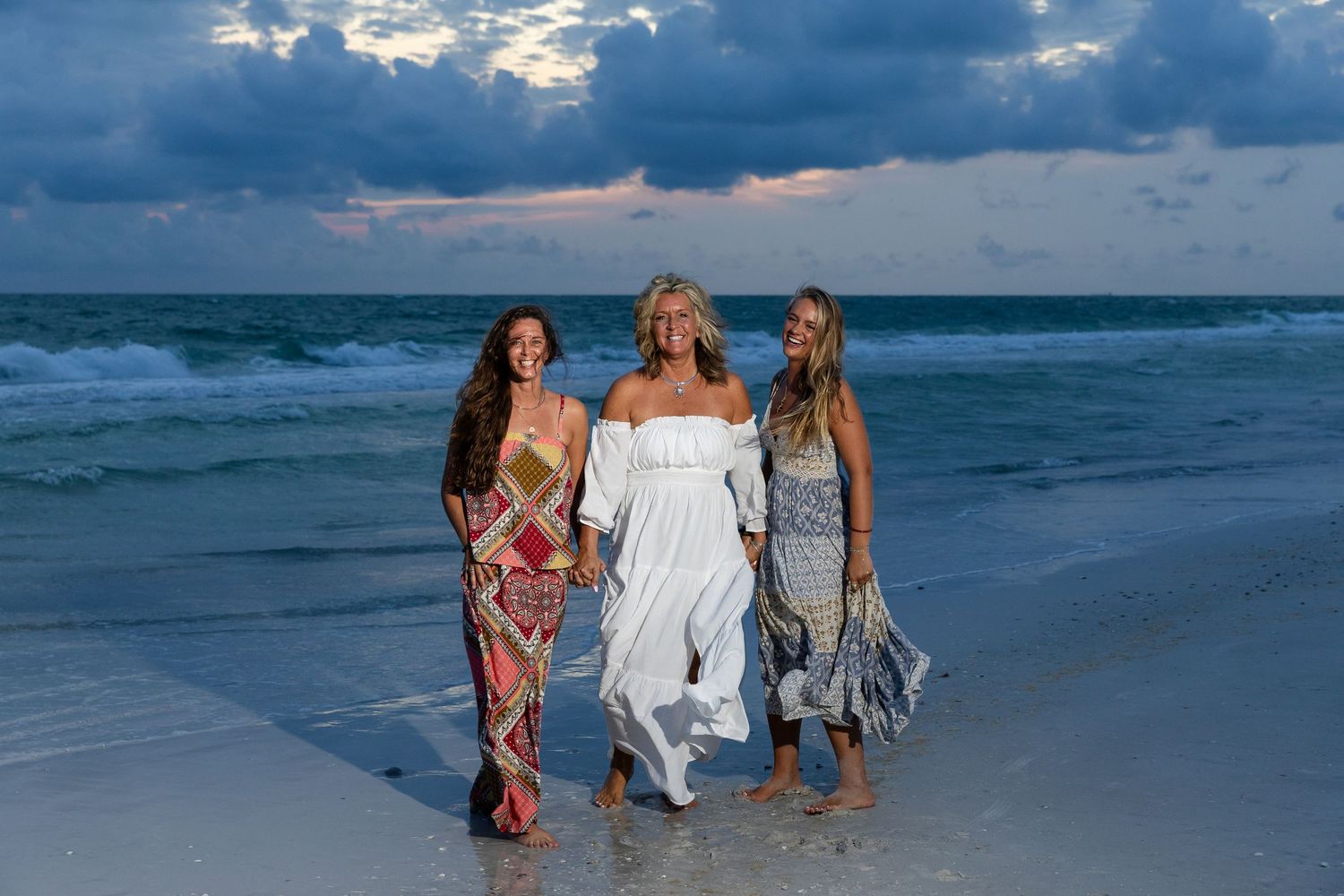 Three women walking on a beach at sunset, wearing colorful summer dresses, with ocean waves and dramatic cloudy sky.