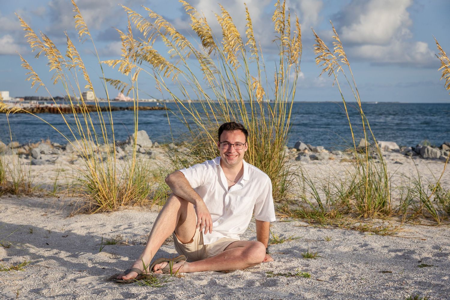 Smiling young man in white shirt sitting on sandy beach with sea oats, rocks, and ocean in background.