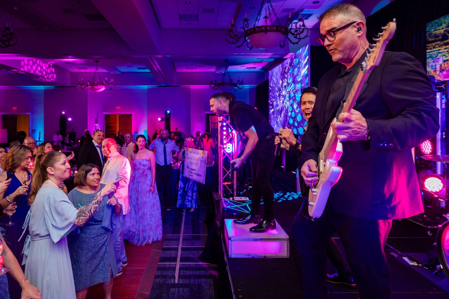 Live band guitarist performing on stage at an elegant indoor event with dancing guests.