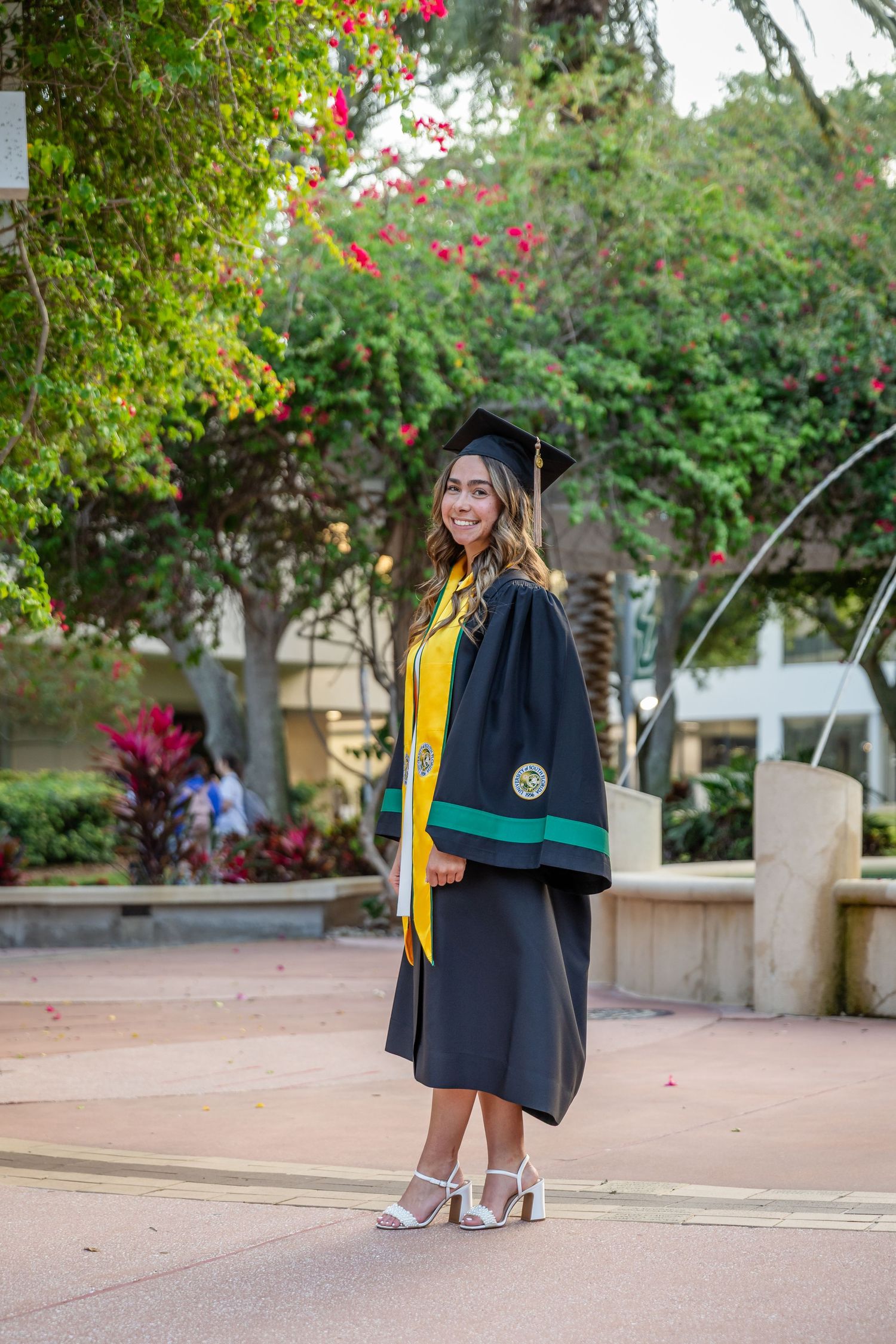 Smiling graduate in black cap and gown with yellow sash poses outdoors near flowering trees and fountain.