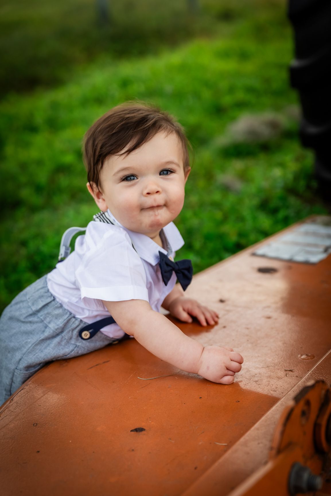 Adorable baby boy in white shirt, bow tie, and suspenders smiles while leaning on a rustic surface outdoors.