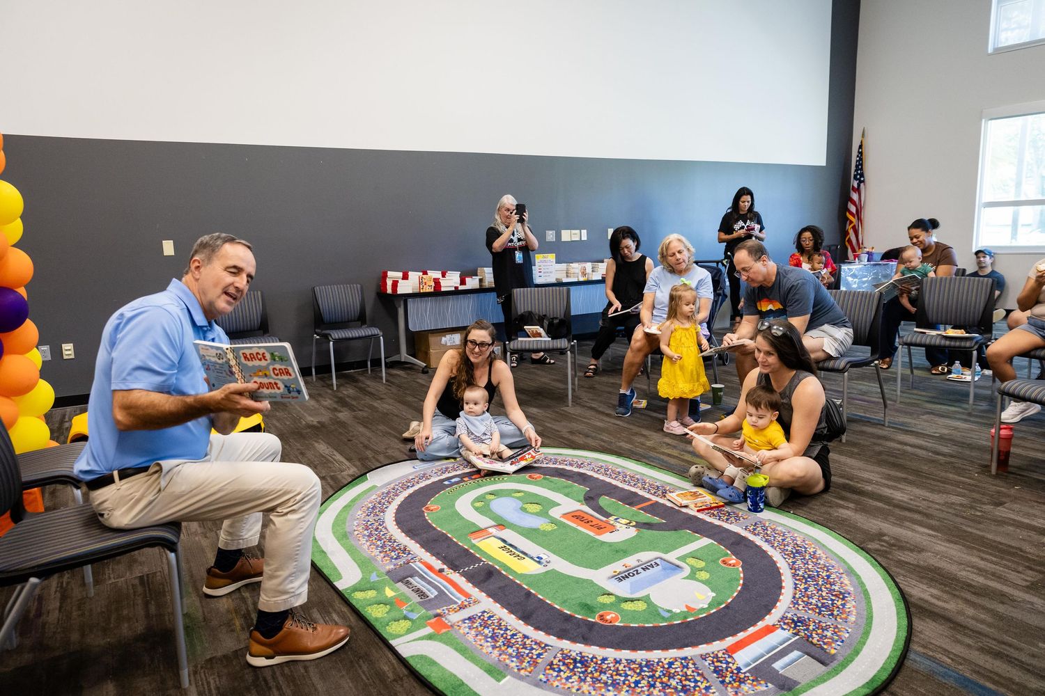 Adults and children sit on a colorful race track rug during a storytime reading session in a bright community room.