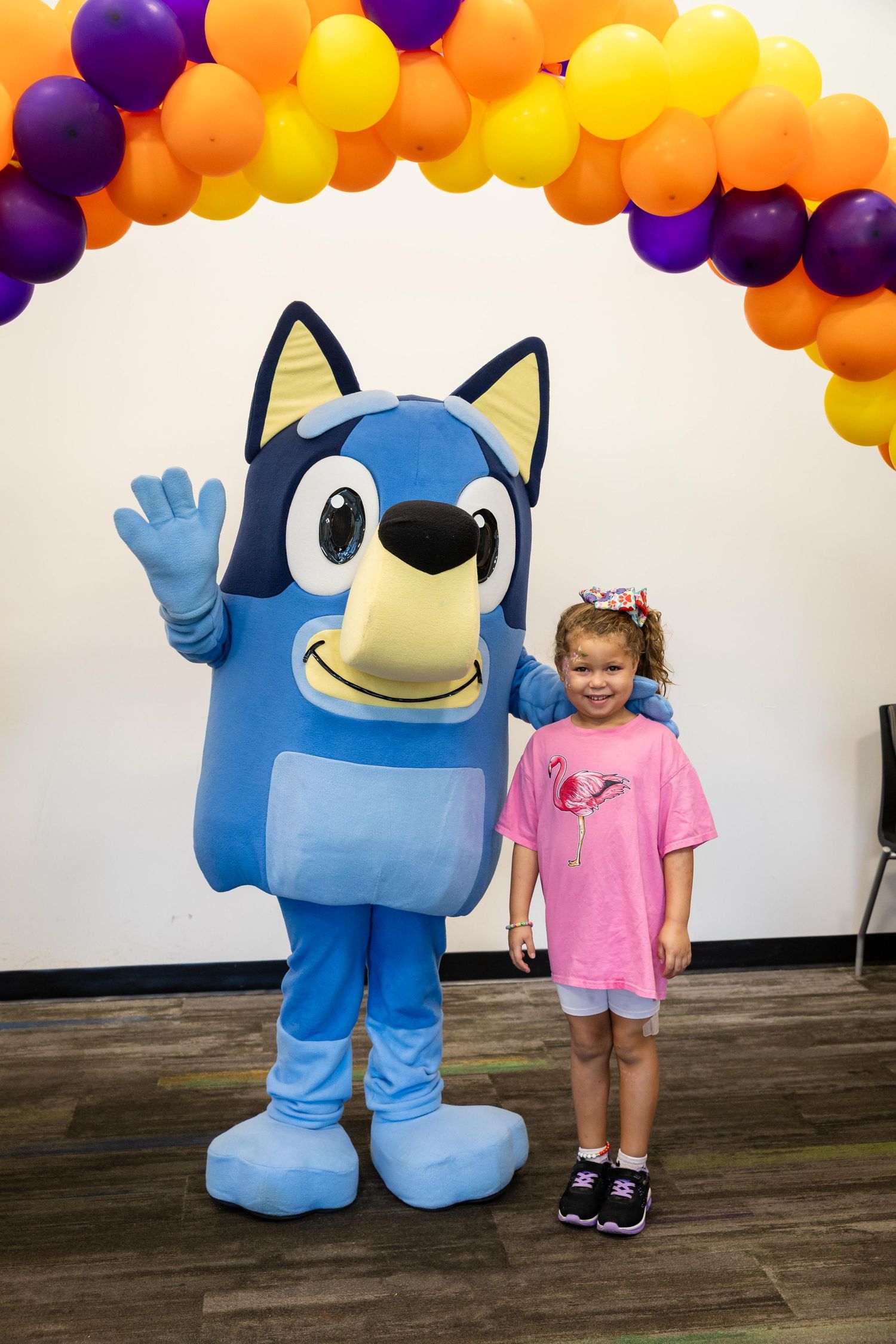 A young girl poses with a Bluey mascot costume character under a colorful balloon arch at a fun event.