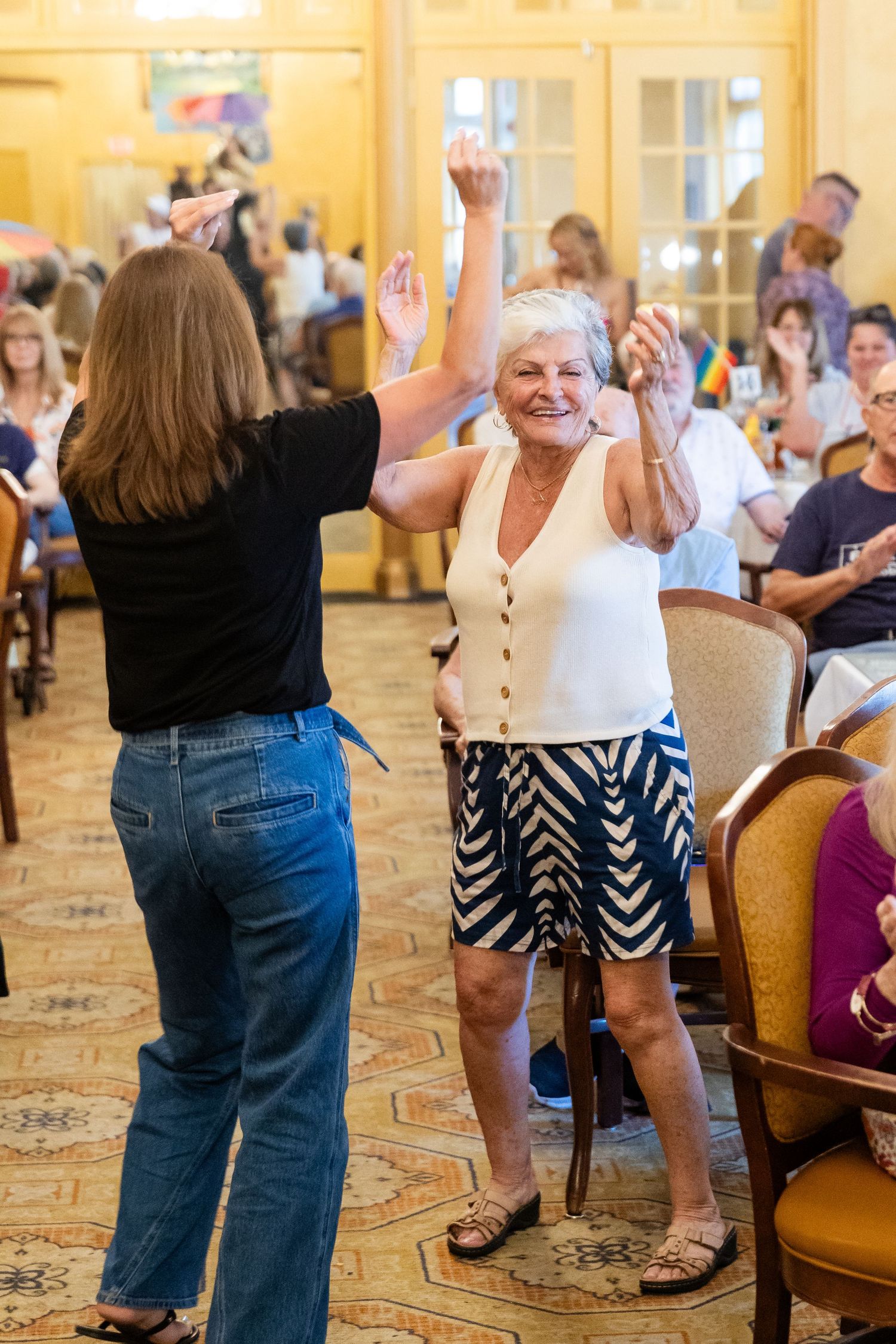 Two women dancing together with raised hands at a lively senior community social event indoors.