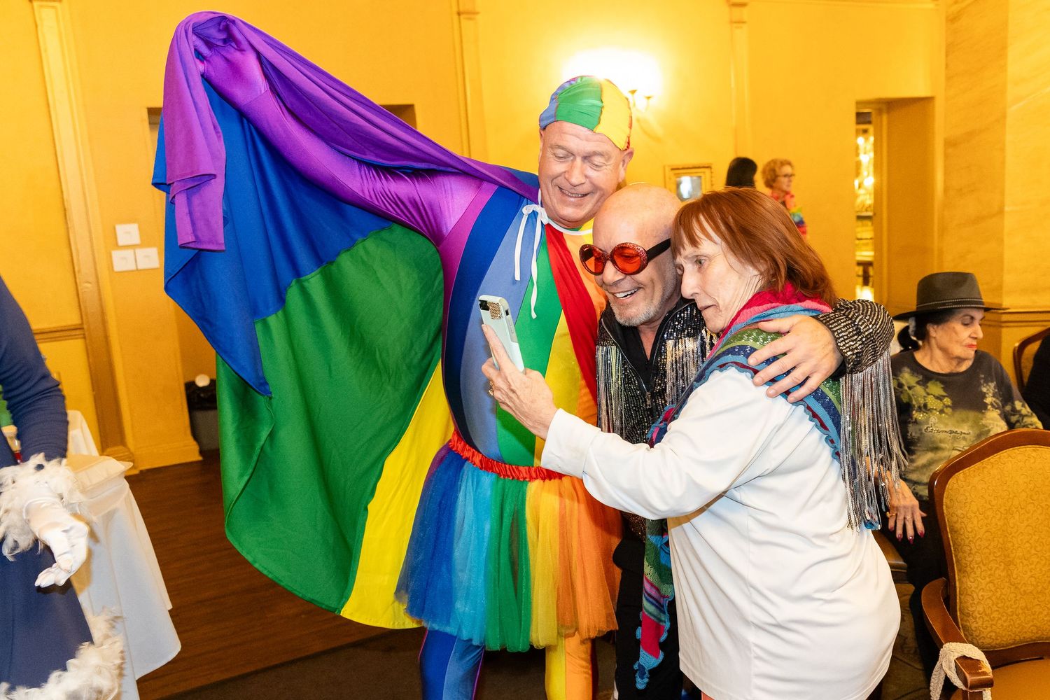 Person in colorful rainbow costume poses for selfie with attendee at an indoor Pride celebration event.