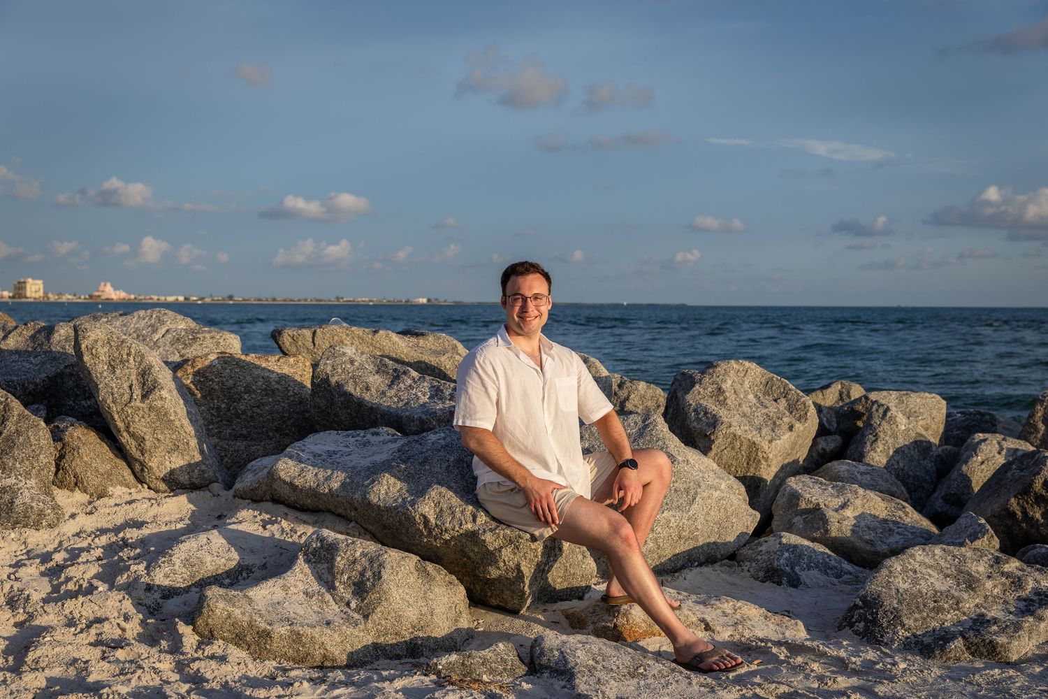 Man sitting on rocky jetty by the ocean in white shirt and shorts on a sunny day.