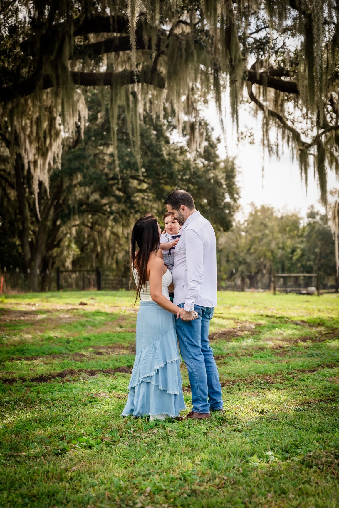 A couple shares a tender moment holding their baby under Spanish moss-covered trees in a green field.