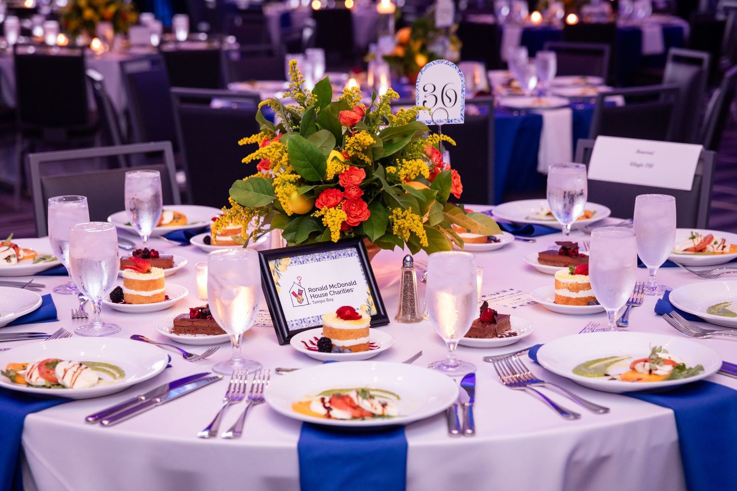 Elegant gala dinner table setting with floral centerpiece, white linens, blue accents, and plated desserts at a formal event.