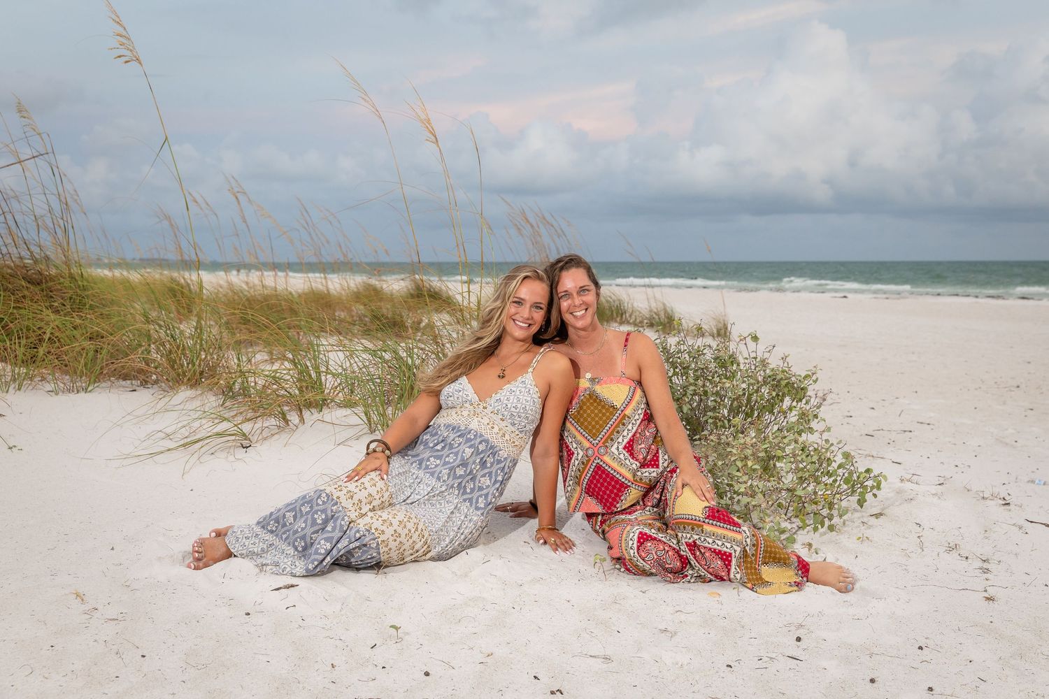 Two women in colorful maxi dresses smile together on a white sandy beach with sea oats and ocean backdrop.