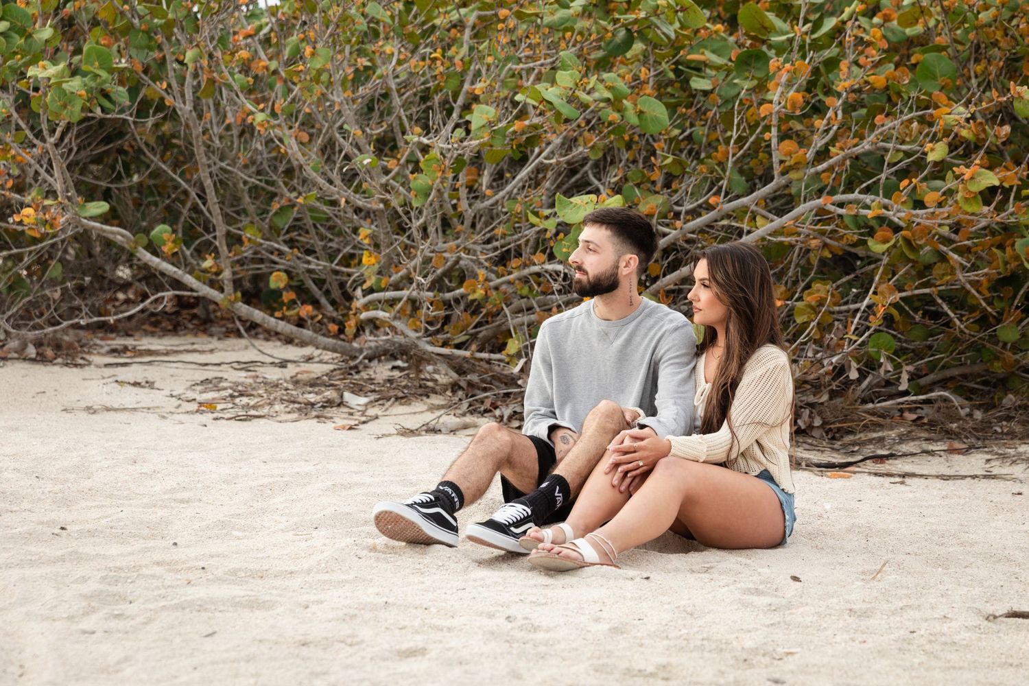 A young couple sits together on sandy beach, relaxing near lush green shrubs, enjoying a peaceful outdoor moment.