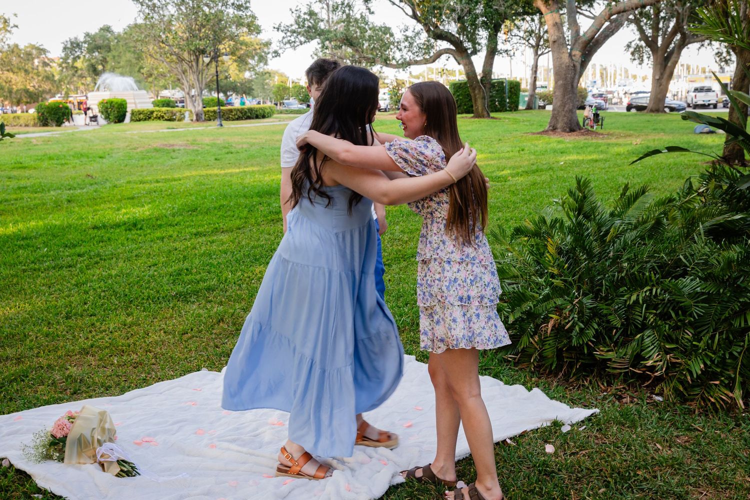 Two young women in summer dresses embrace on a white blanket in a lush green park.