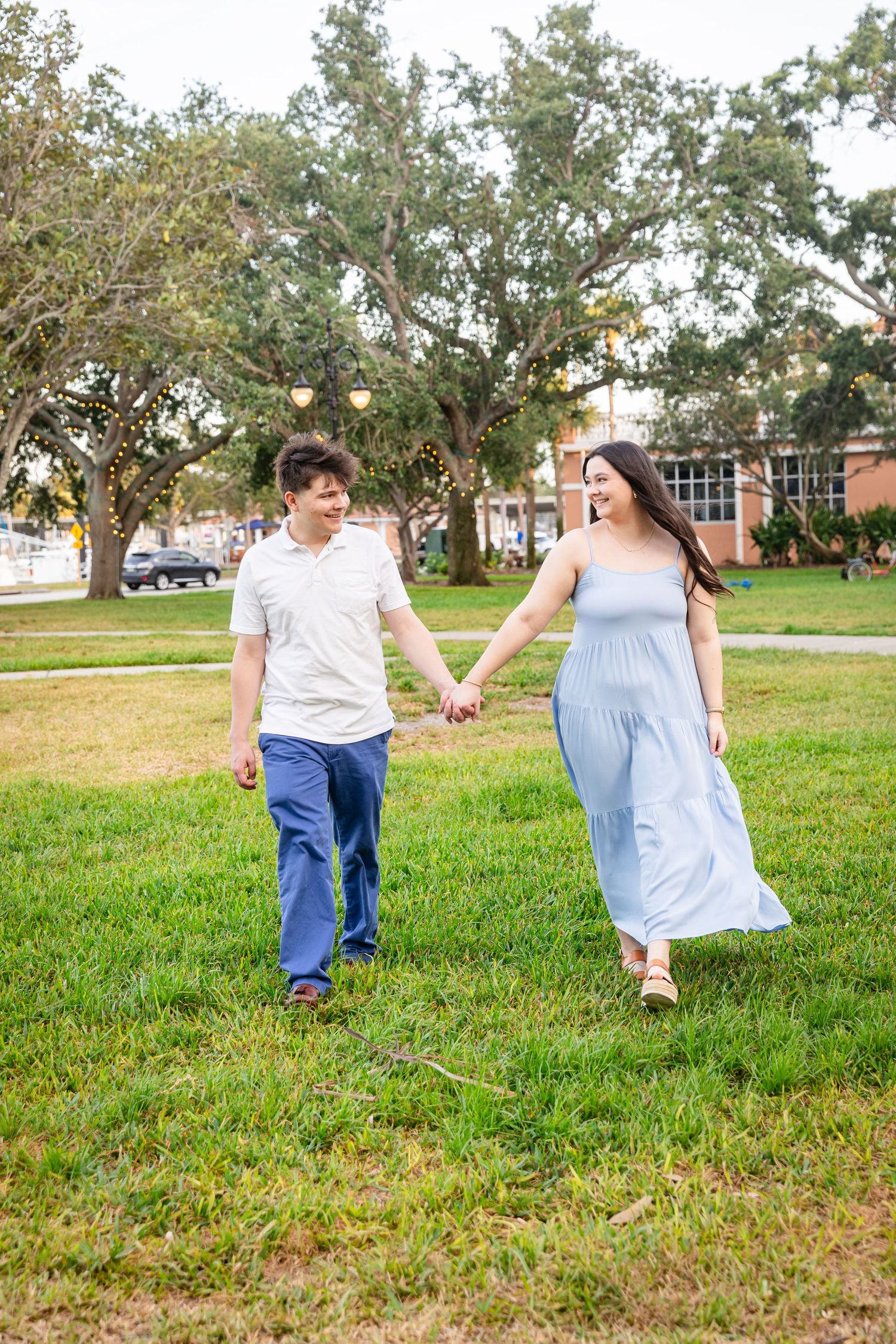 A couple holds hands walking through a green park, the woman wearing a flowing light blue maternity dress.