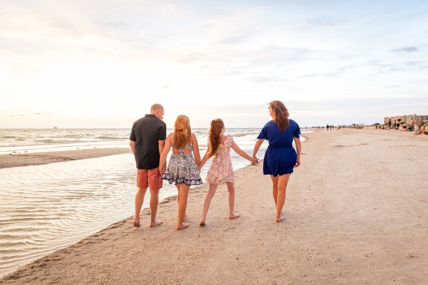 Family of four walking hand-in-hand along a sandy beach at sunset, wearing casual summer outfits.