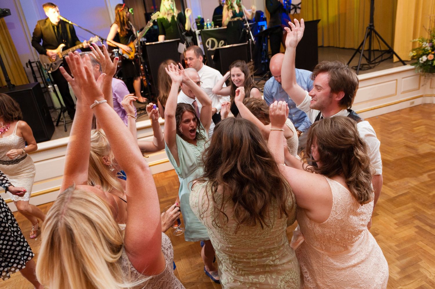 Wedding guests celebrate on dance floor with hands raised at lively reception party.