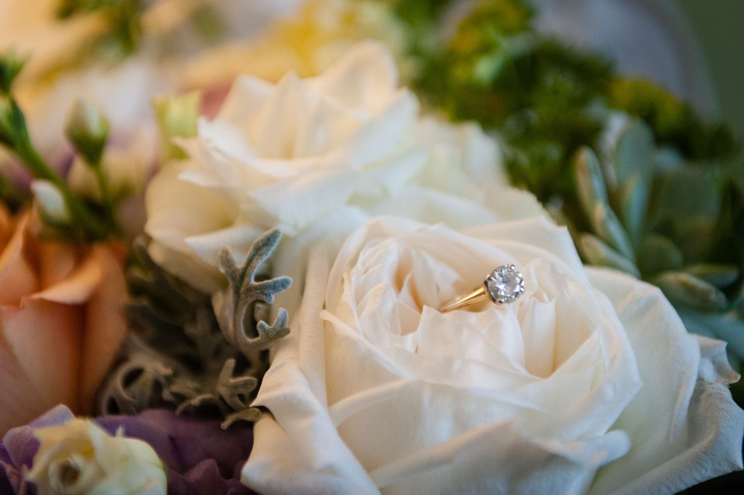 Gold diamond engagement ring resting elegantly among soft white roses in a bridal bouquet.