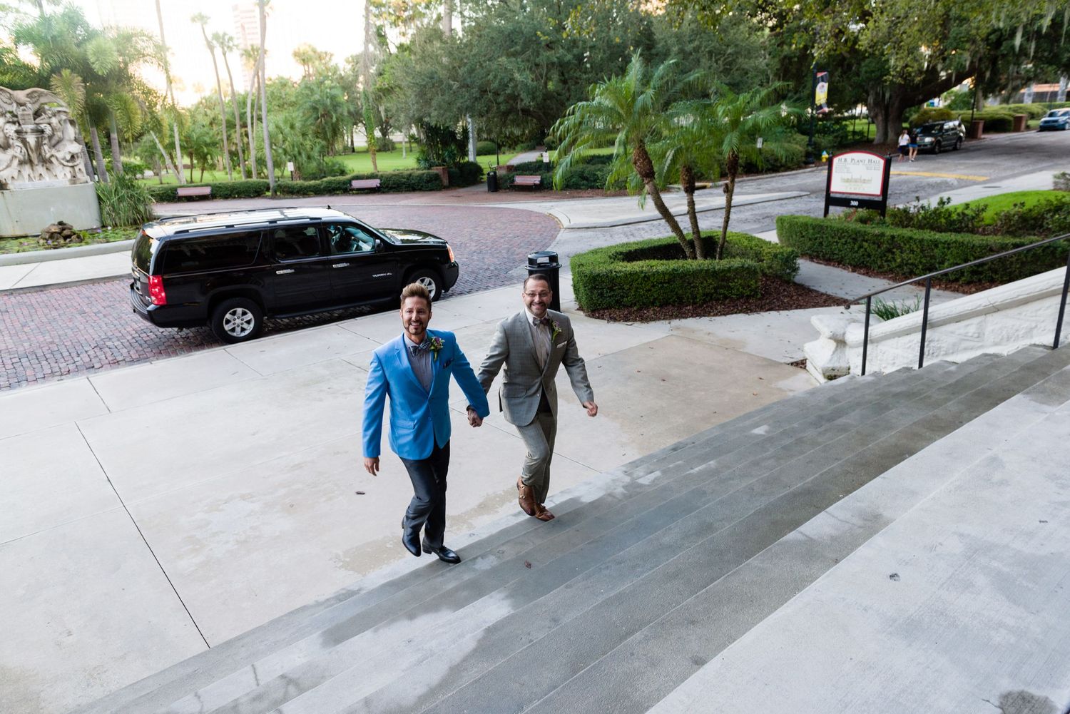 Two men holding hands walk up steps outside a tropical venue with palm trees and a black van.