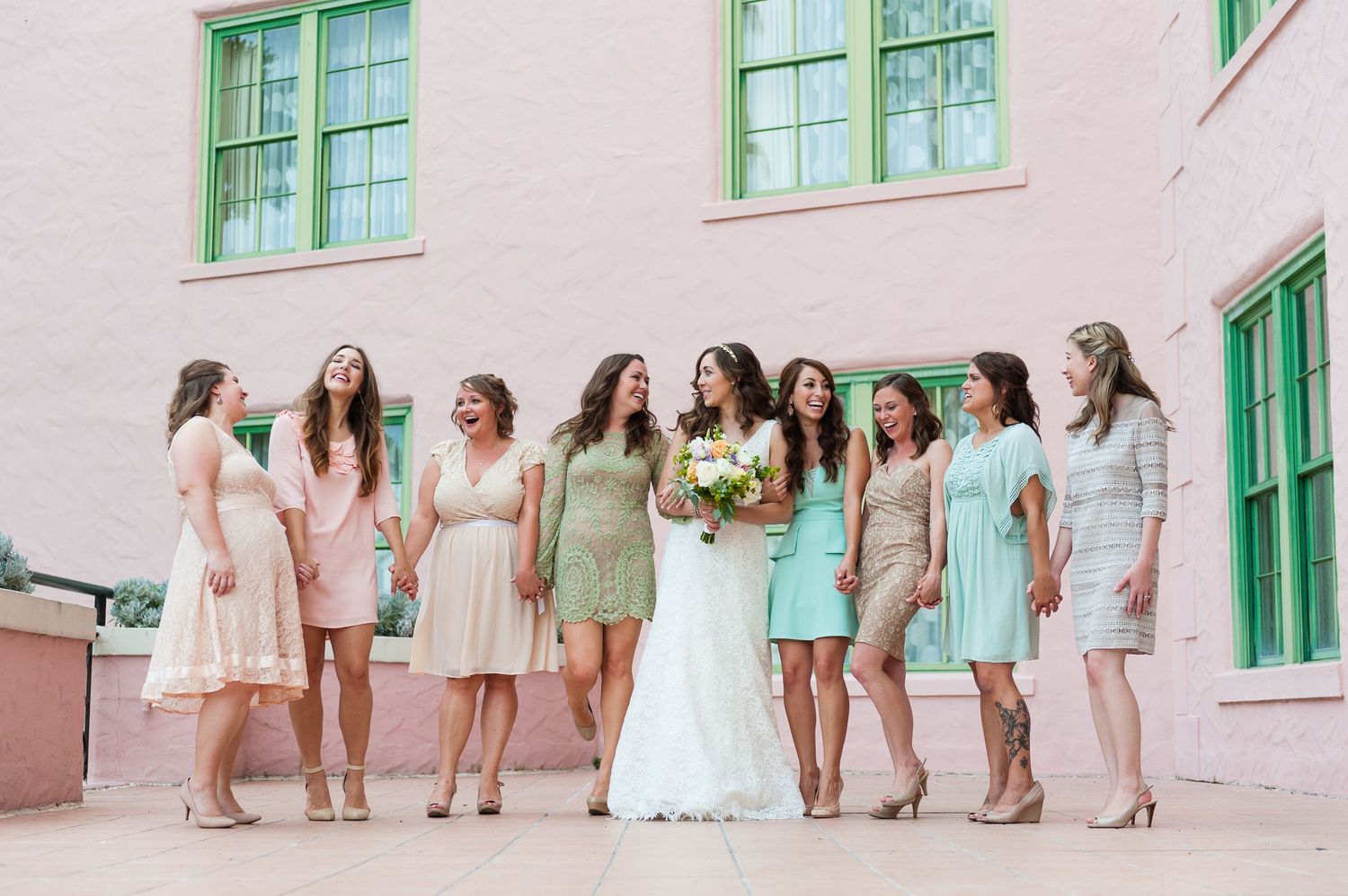 Bride and bridesmaids in mismatched pastel dresses laughing together outside a pink building.