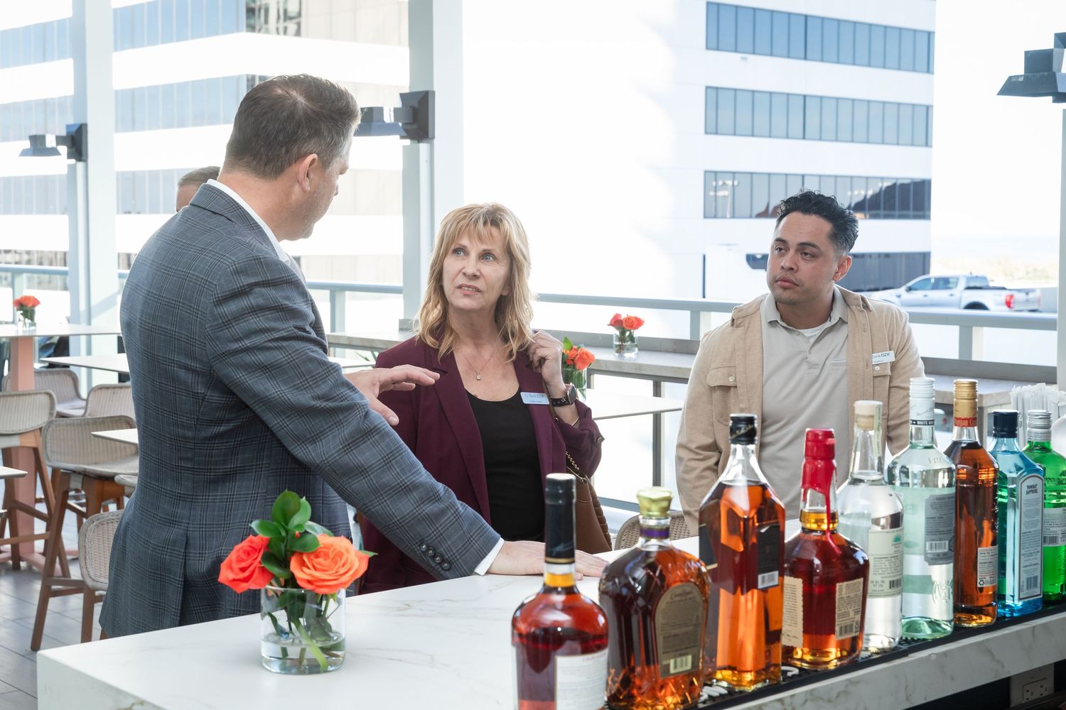 Three professionals networking at a rooftop bar with liquor bottles, engaged in conversation at a modern venue.