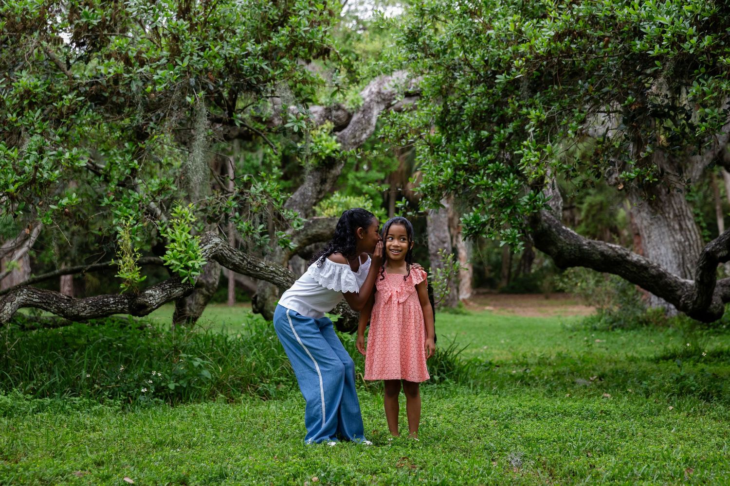 Two girls sharing a secret moment in a lush green forest park with ancient twisted trees.