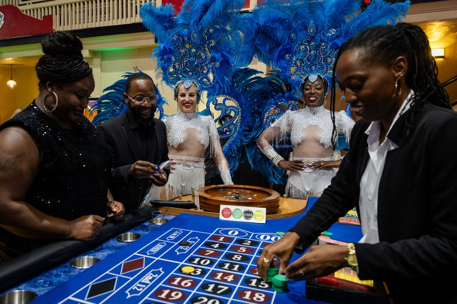 People playing roulette at a casino table with showgirls in blue feathered costumes standing behind them.