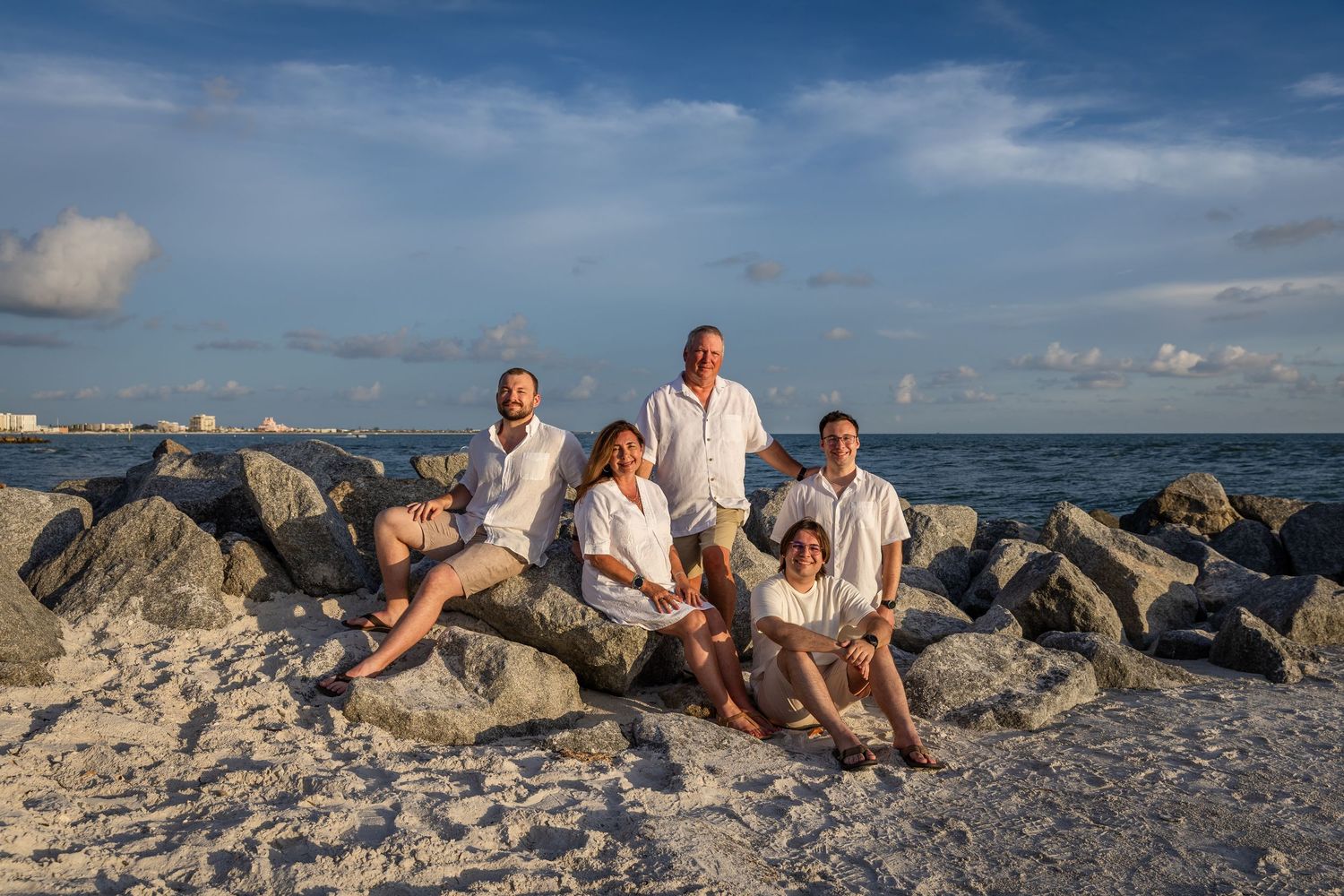Family of five posing on rocky beach shoreline with blue ocean and cloudy sky backdrop during golden hour.