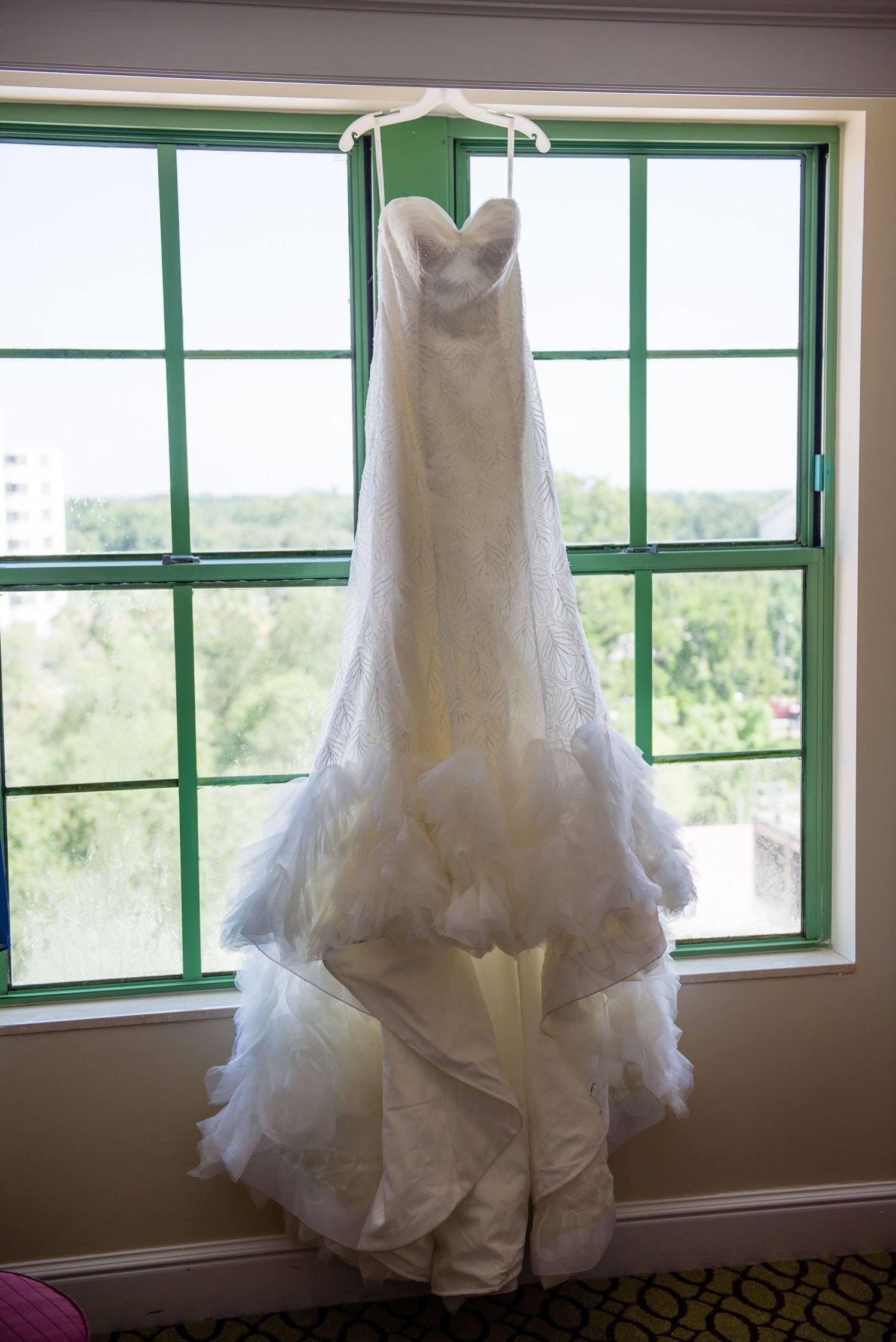 Elegant white lace mermaid wedding dress hanging on a hanger in front of a green-framed window.