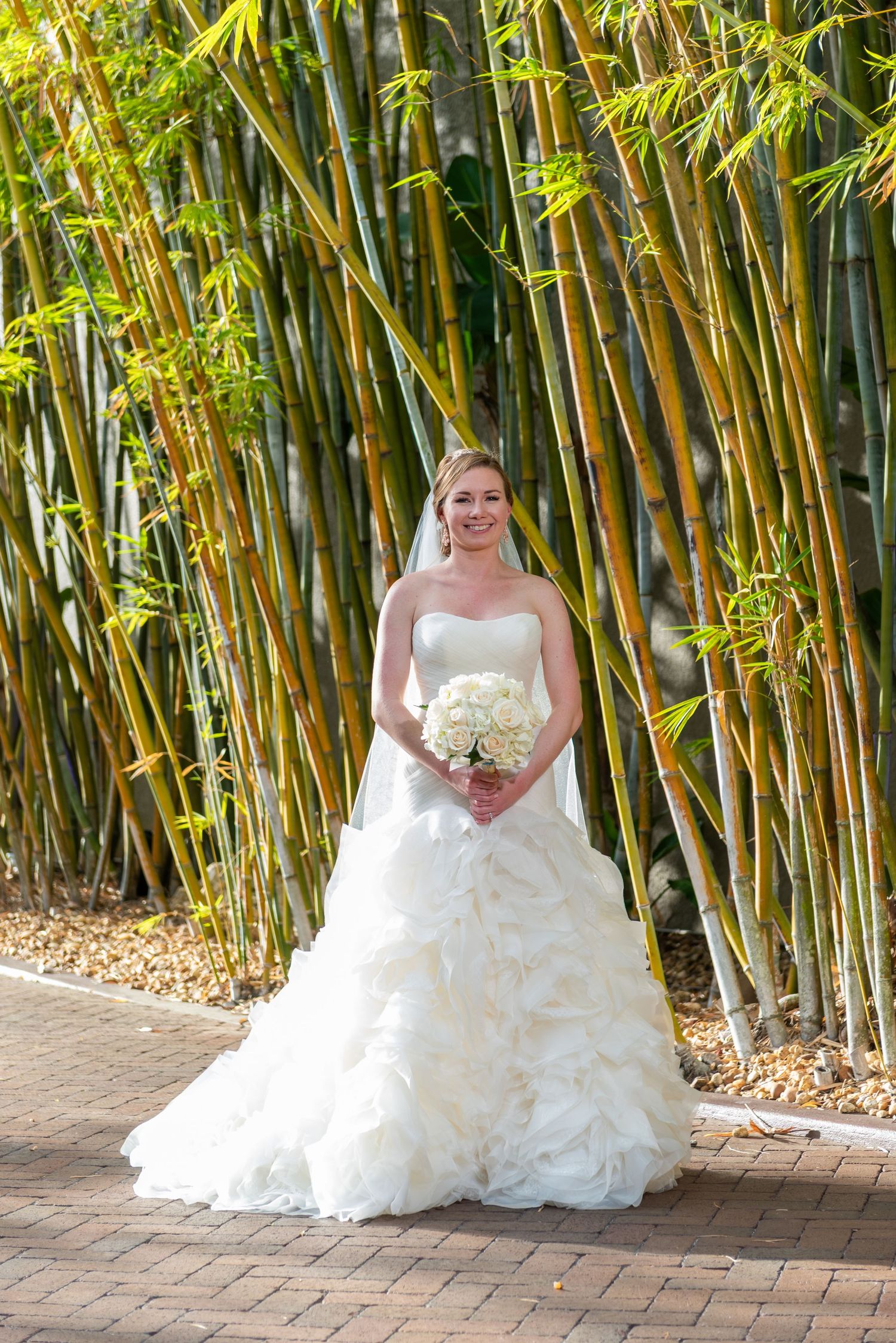 Bride in strapless ruffled white ball gown holds white bouquet while posing in front of tall bamboo trees.