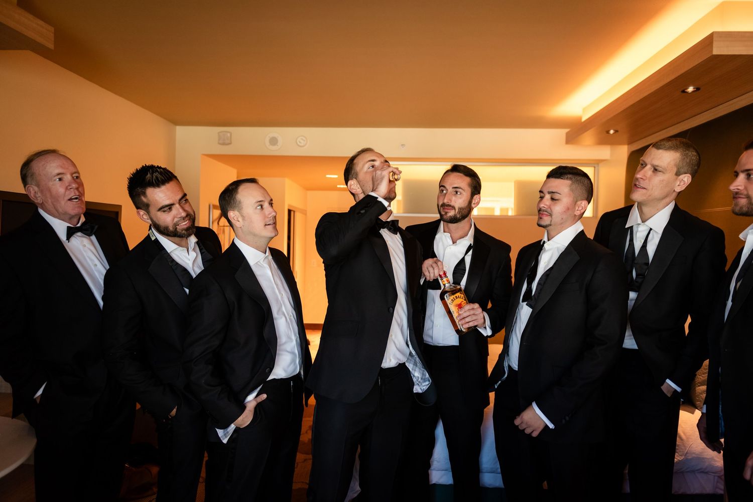 Groomsmen in black tuxedos laughing and celebrating together in a hotel room before a wedding ceremony.