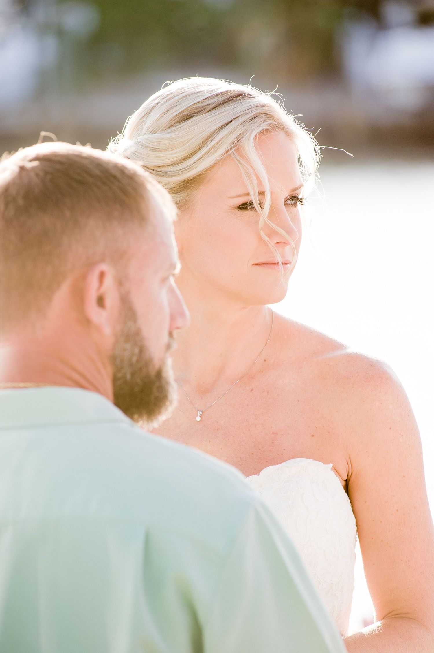 Blonde bride in strapless wedding gown gazes lovingly at bearded groom during an outdoor beach ceremony.