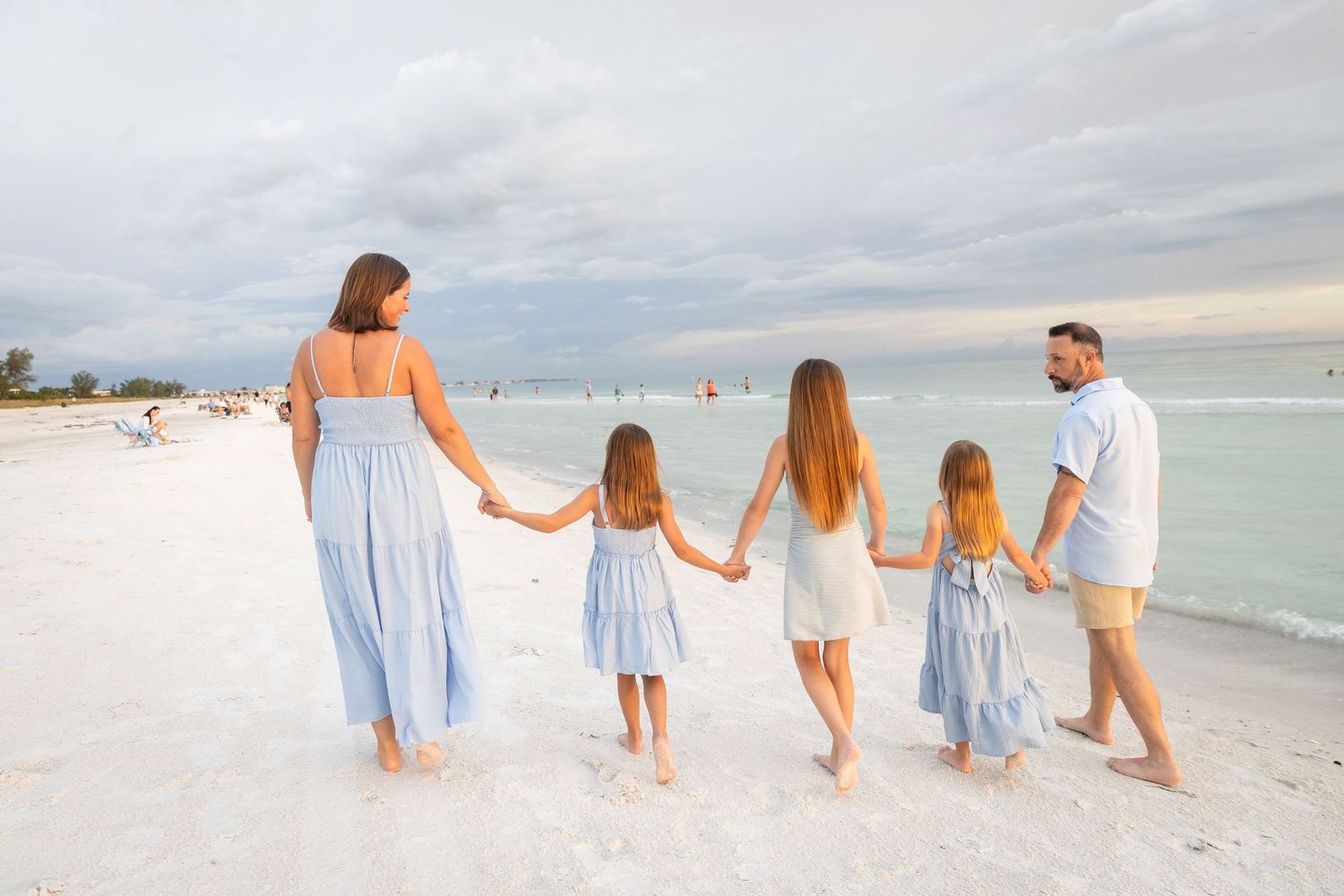 Family of five walking hand-in-hand on a white sand beach at sunset, dressed in coordinating blue and white outfits.