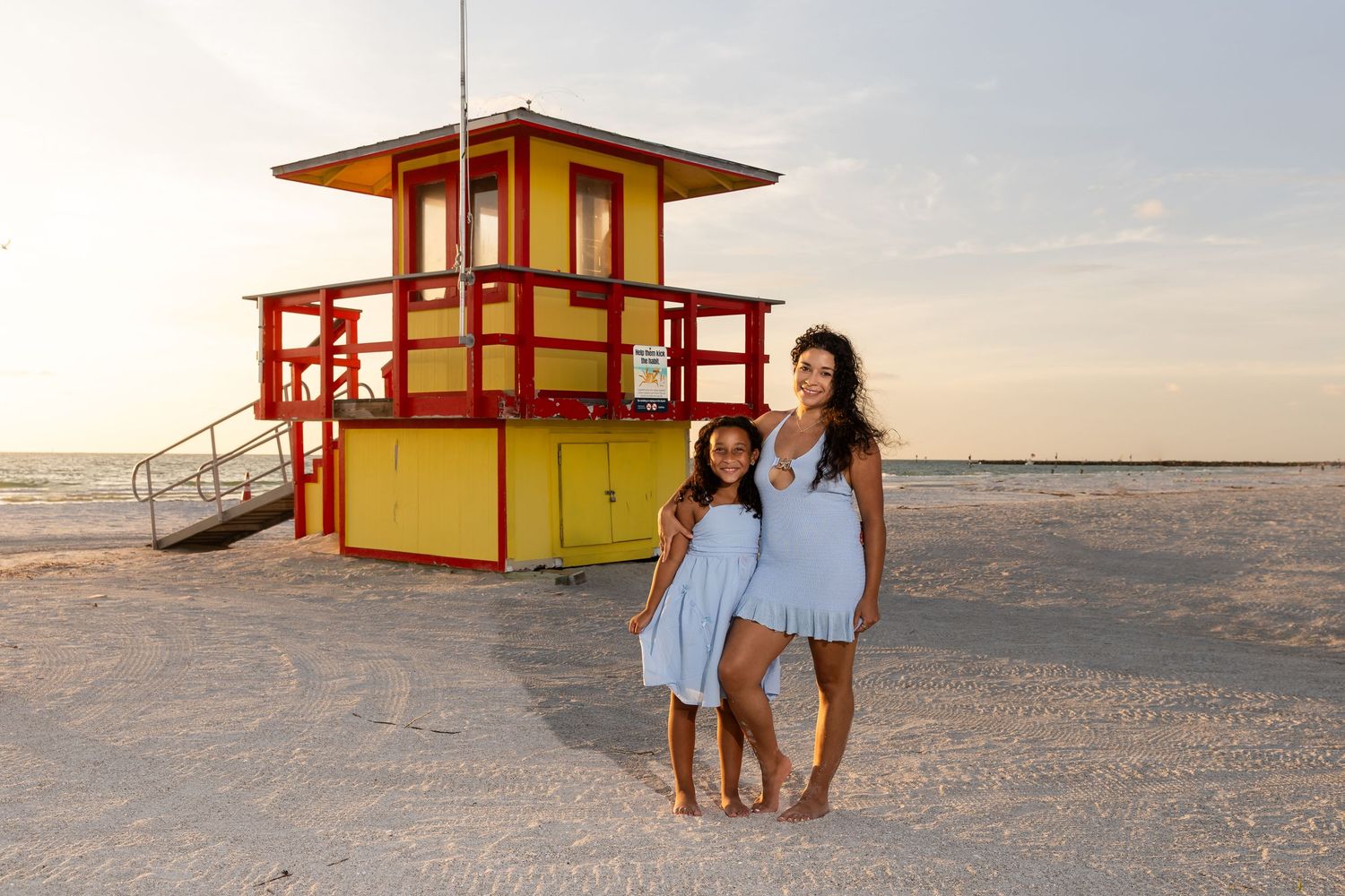 Mother and daughter in blue dresses pose in front of a colorful red and yellow lifeguard stand on a sandy beach.