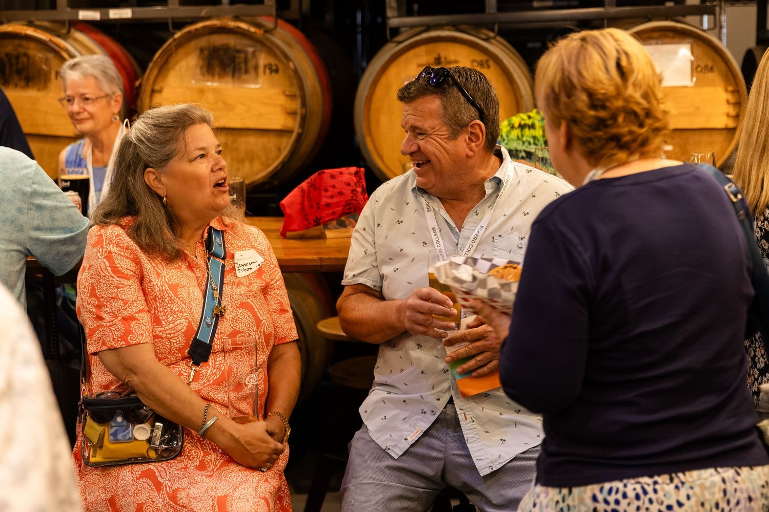 People socializing at a winery with wooden barrels in the background during a wine tasting event.