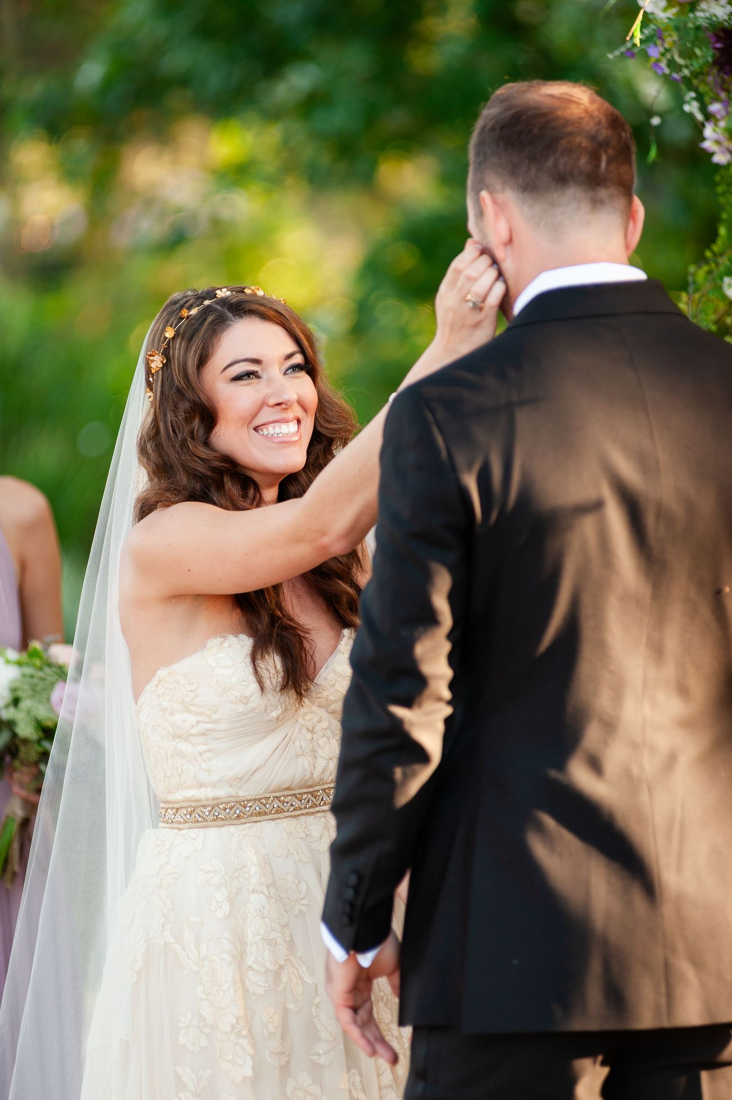 Radiant bride smiles and wipes tear from groom's cheek during emotional outdoor wedding ceremony.