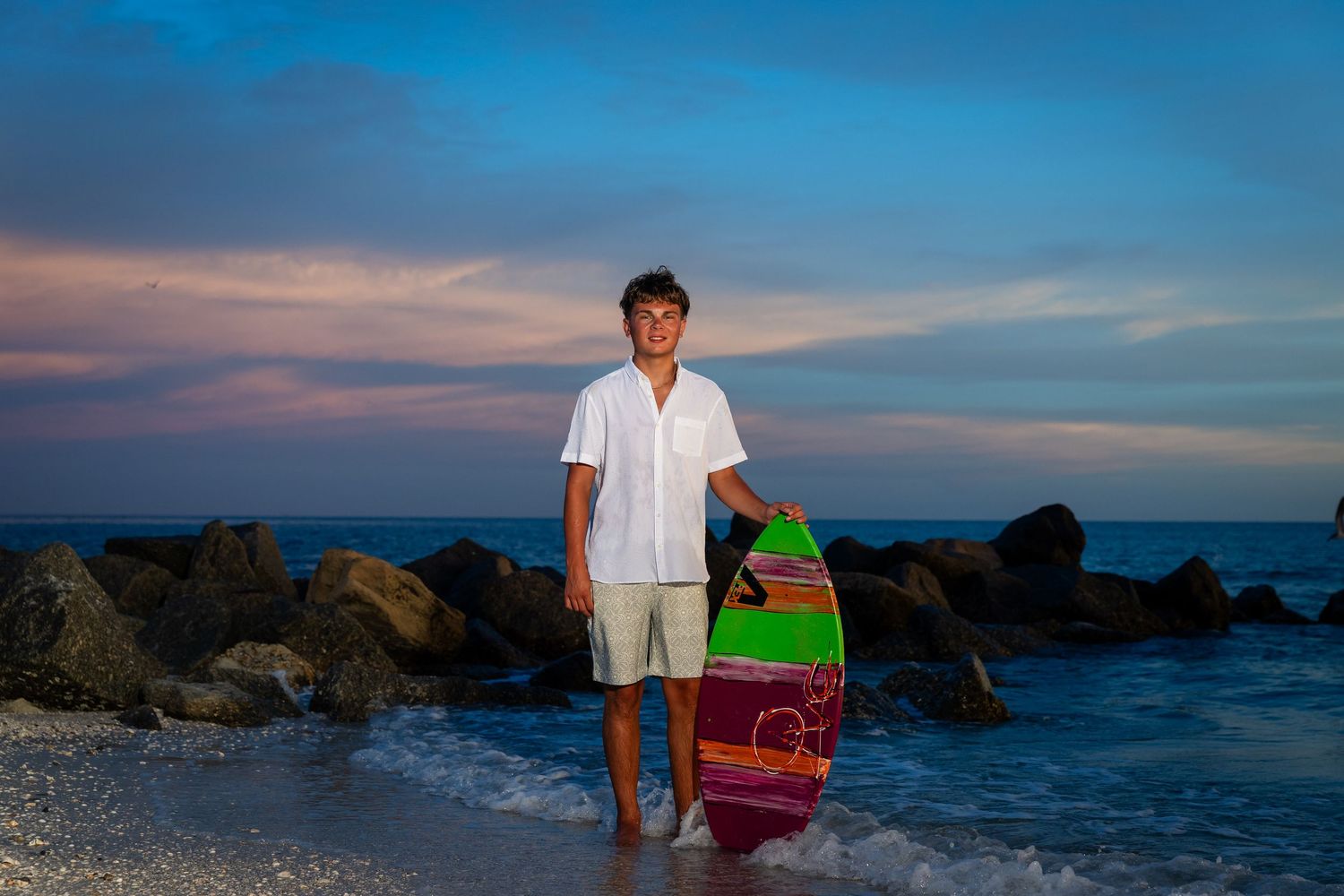 Young man holding a colorful surfboard on a sandy beach at sunset with rocky shoreline and dramatic sky.