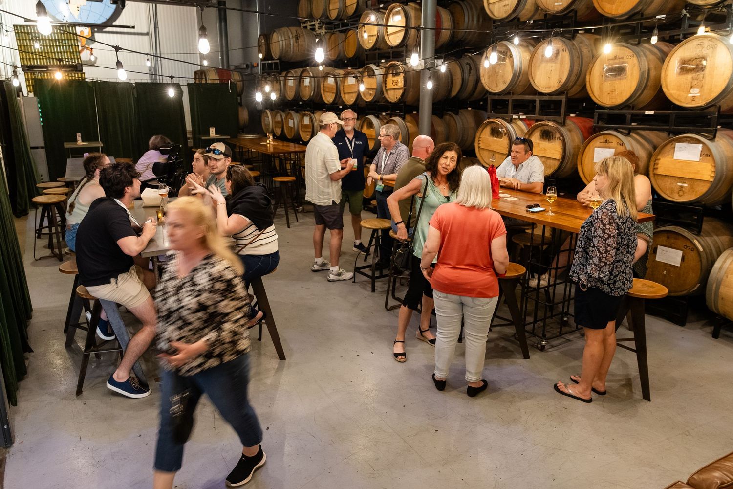 Guests mingle among oak barrels at a lively brewery event with string lights.