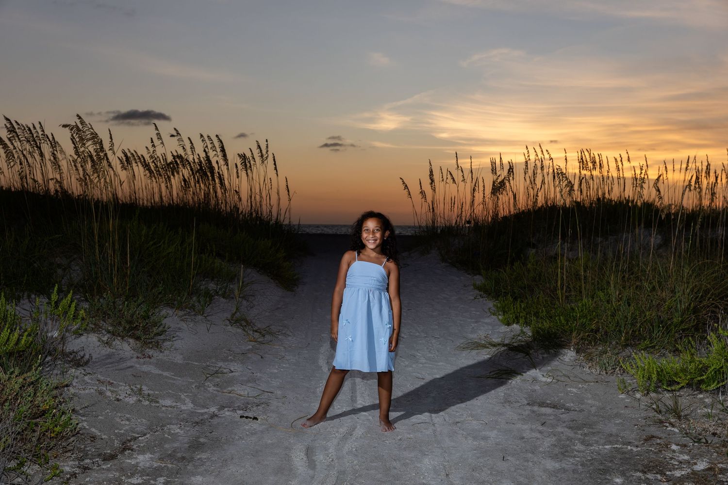 Young girl in blue dress standing on sandy path between sea oats at sunset.