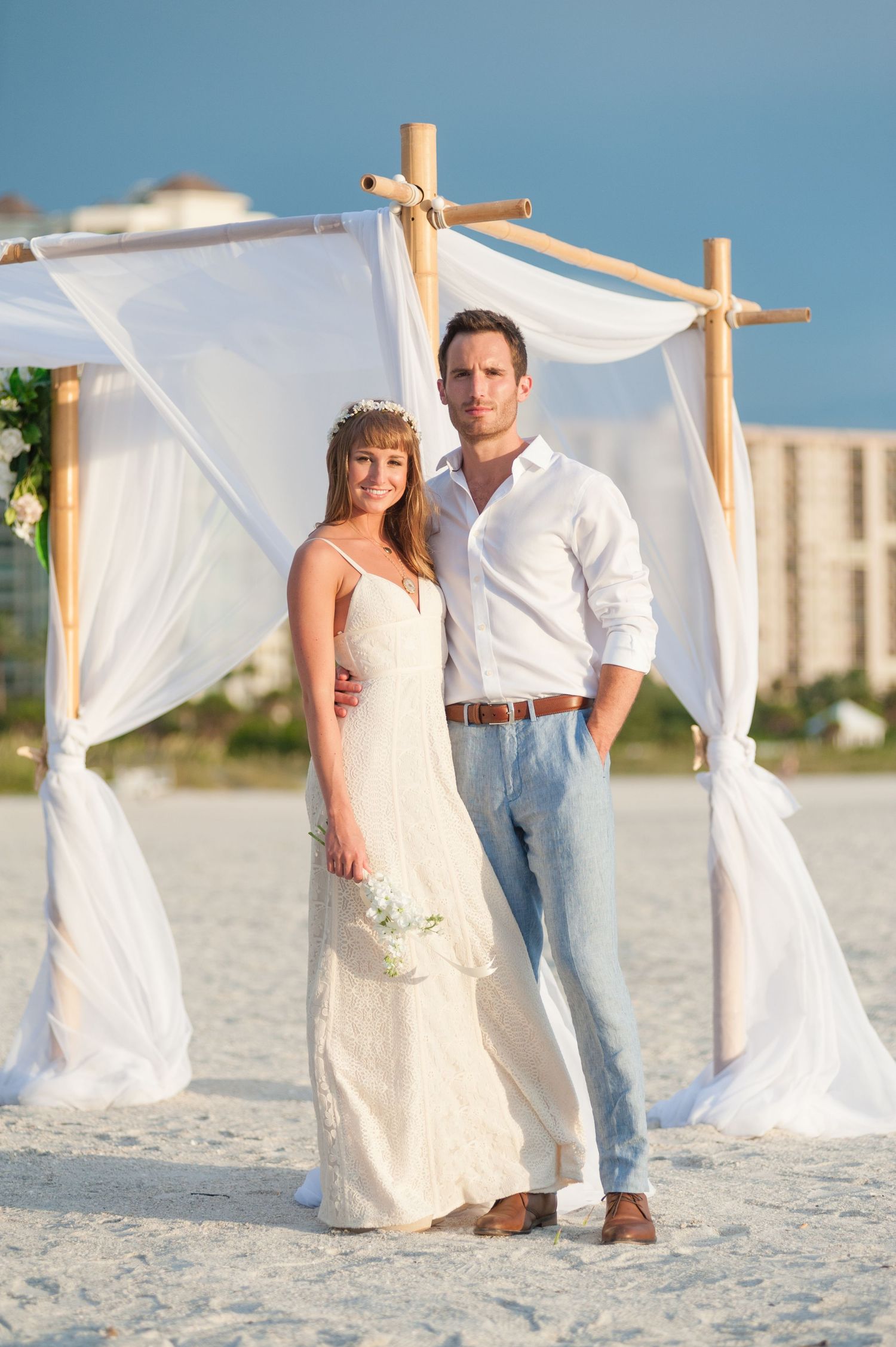 Bride and groom pose under bamboo and white draped arch on sunny beach on Treasure Island, Florida.