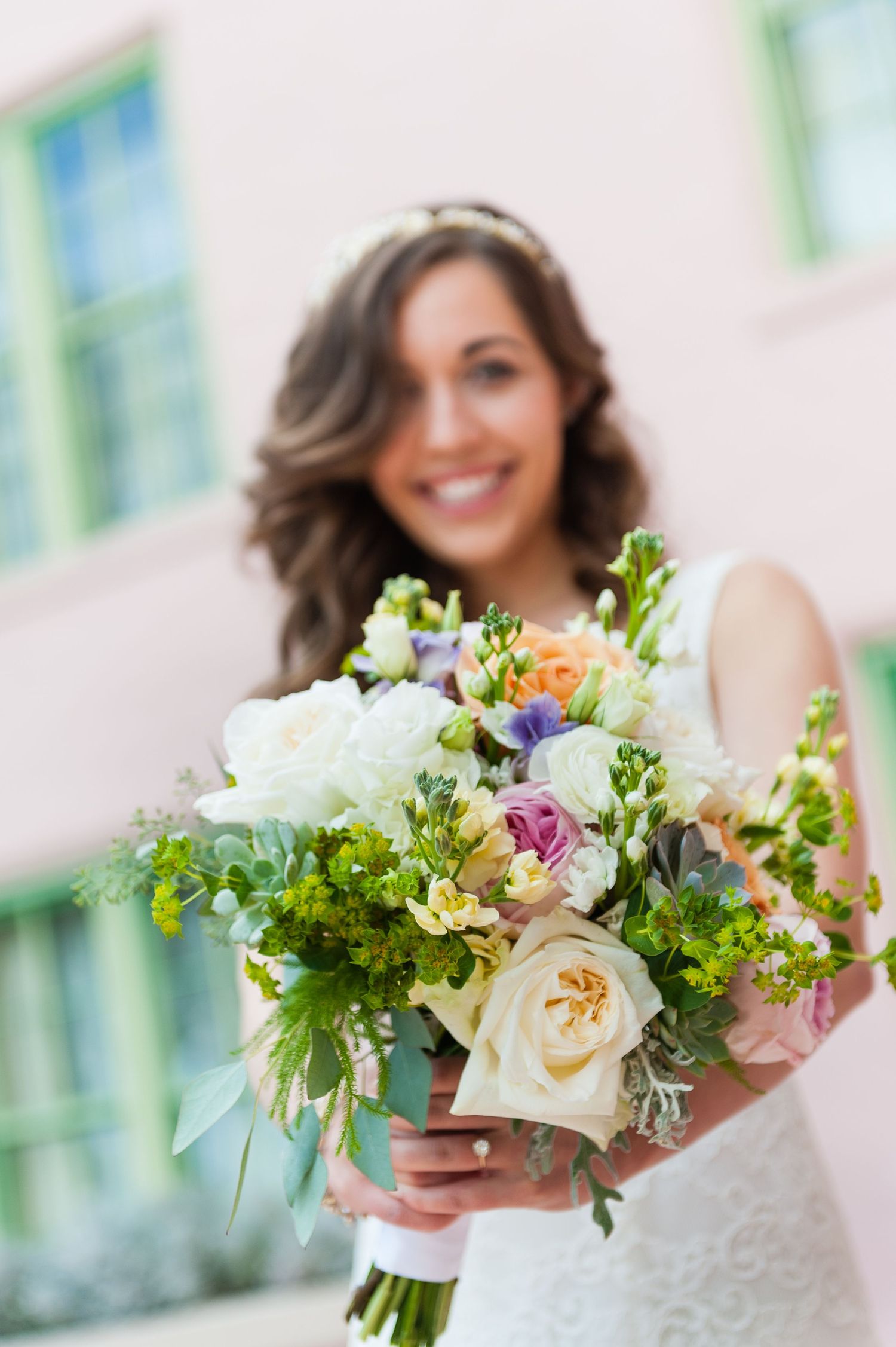 Bride in white lace dress holding a colorful spring wedding bouquet with roses and greenery.