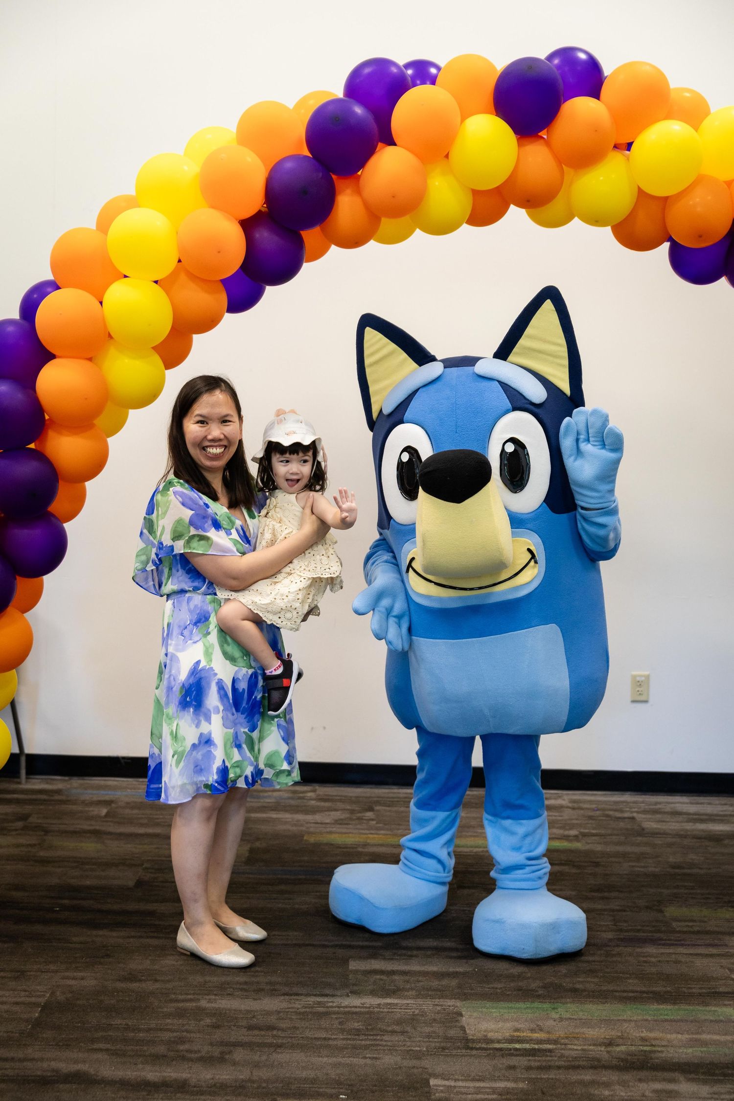 Woman holding toddler poses with Bluey mascot under colorful balloon arch at birthday party.