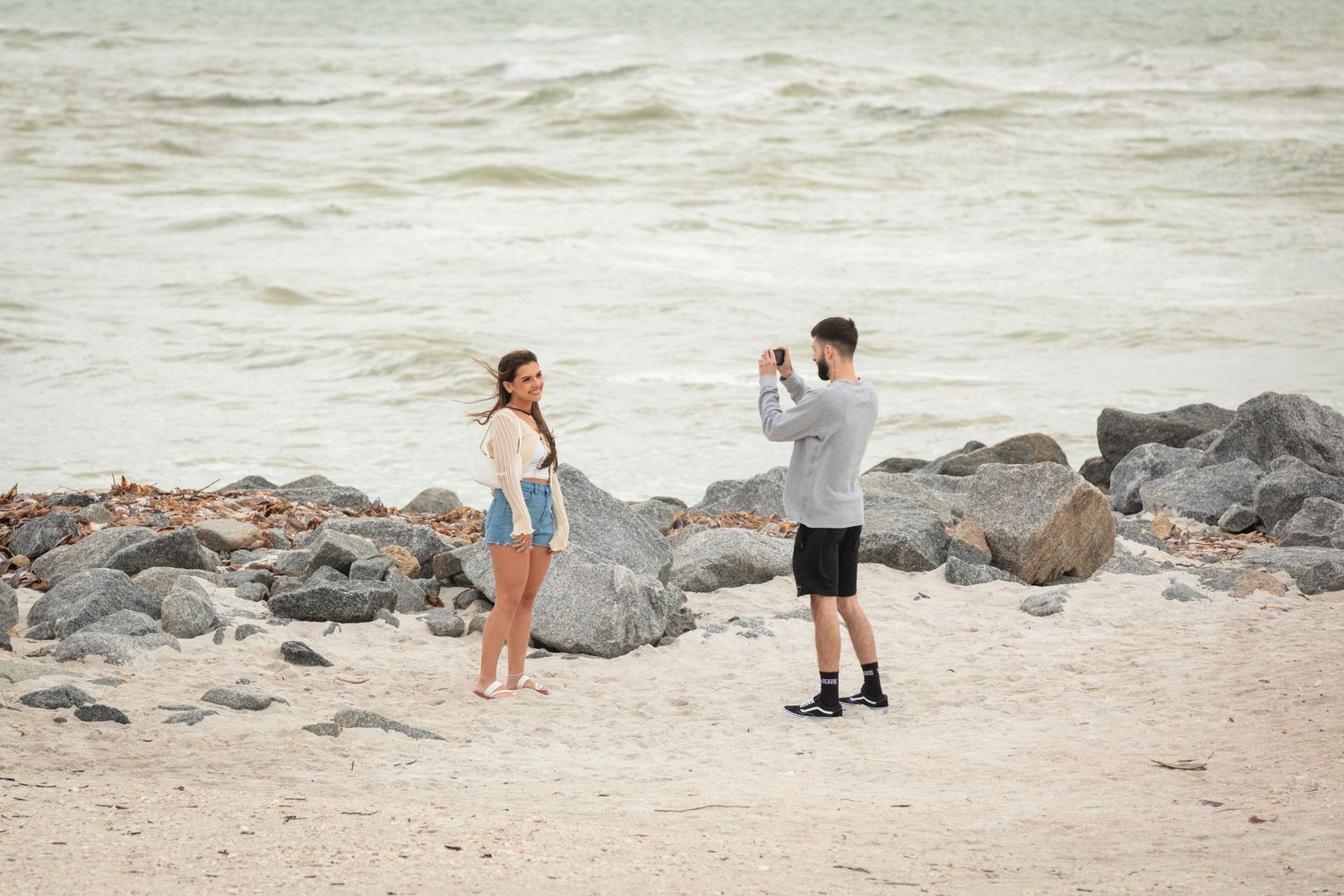 A man photographs a woman posing on a sandy beach with rocky shoreline and ocean waves in the background.