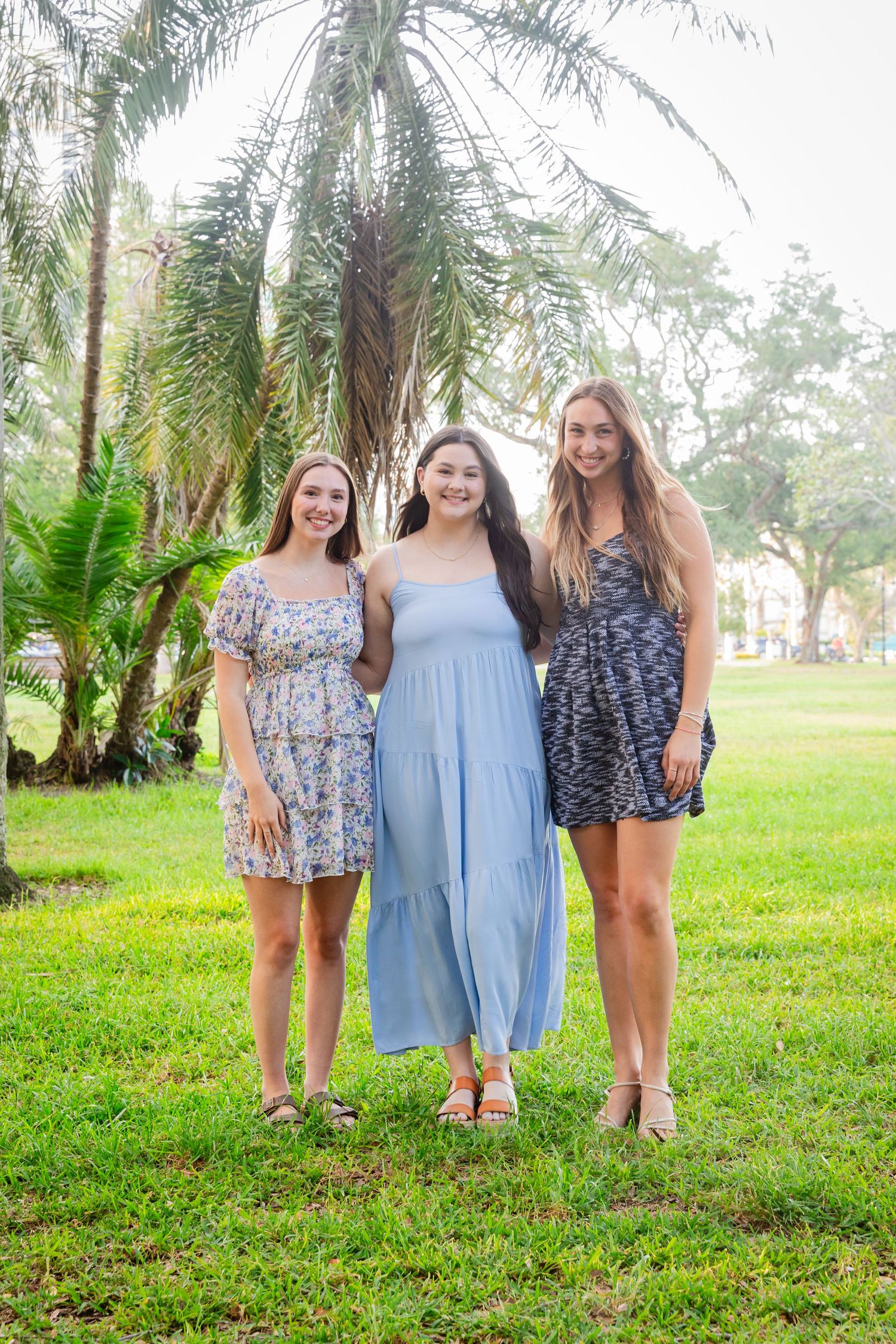 Three smiling young women in floral and blue summer dresses pose together in a tropical park.