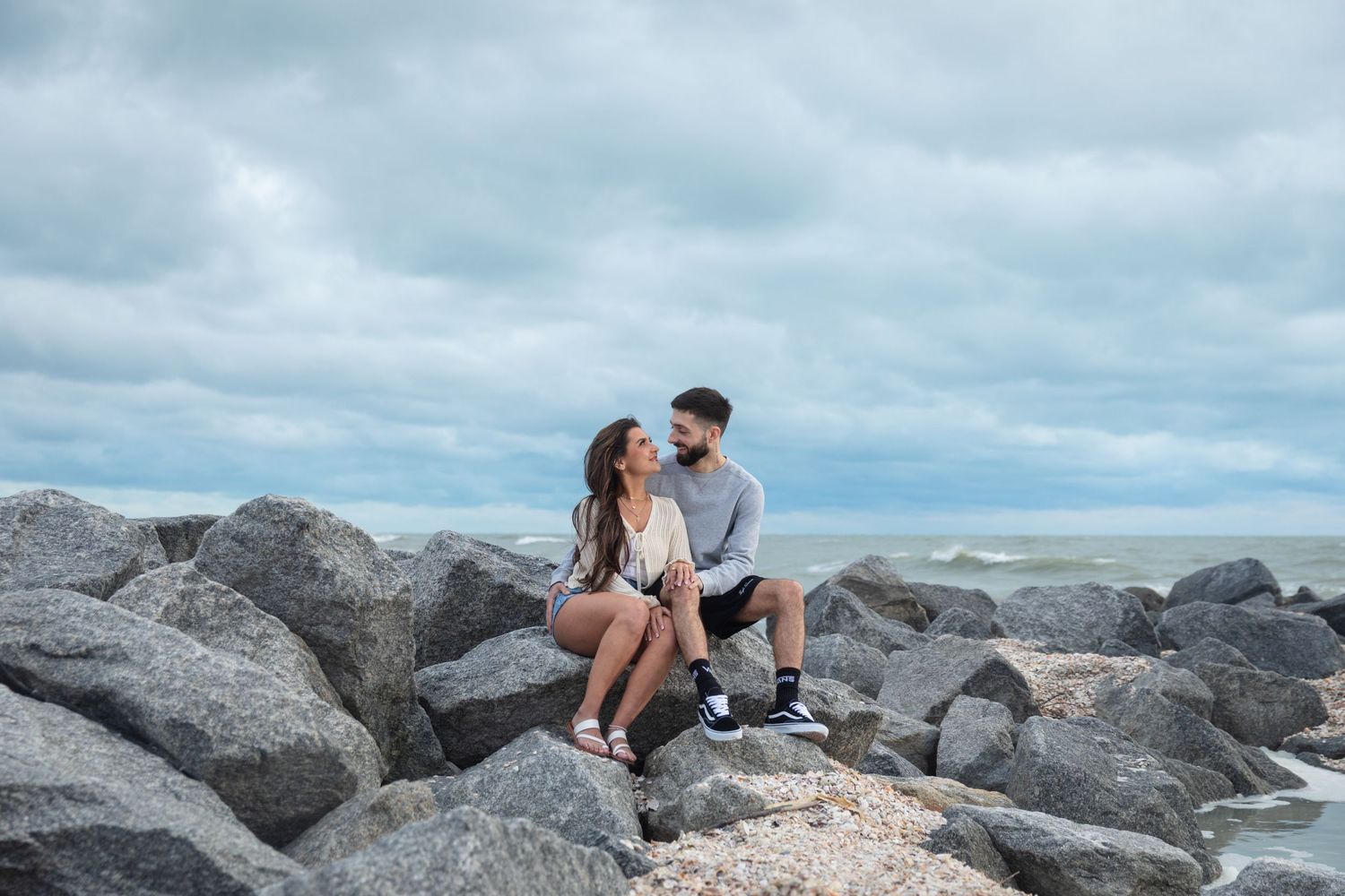 A couple sits on rocky shoreline, gazing at each other with the ocean and cloudy sky in the background.