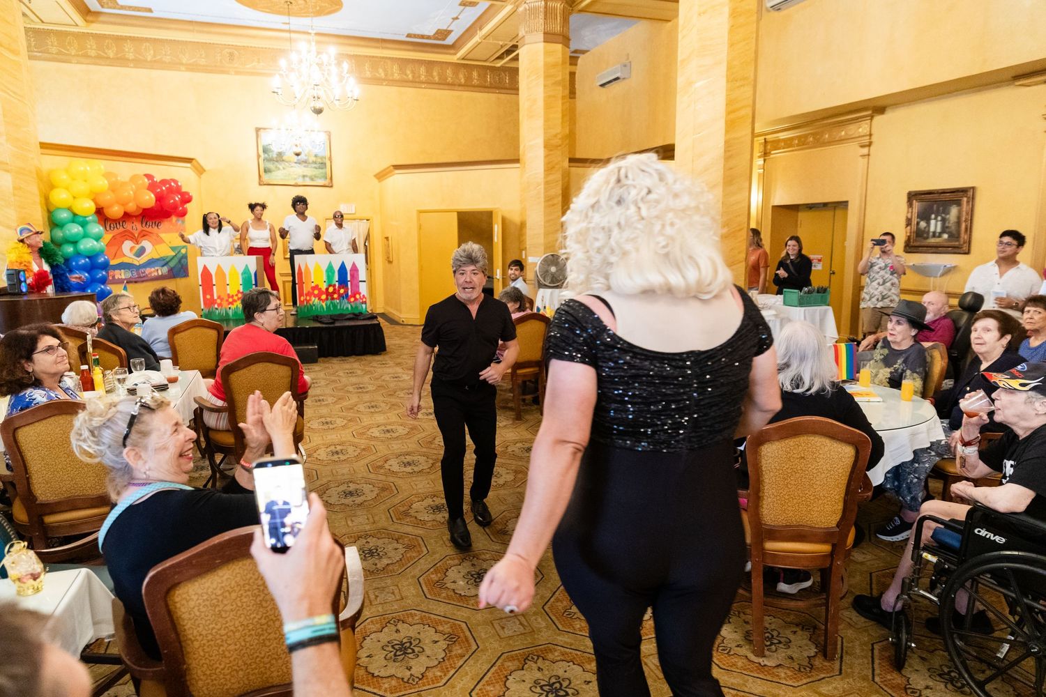 Guests and drag performers mingle on the dance floor at a lively Pride celebration in an ornate ballroom venue.