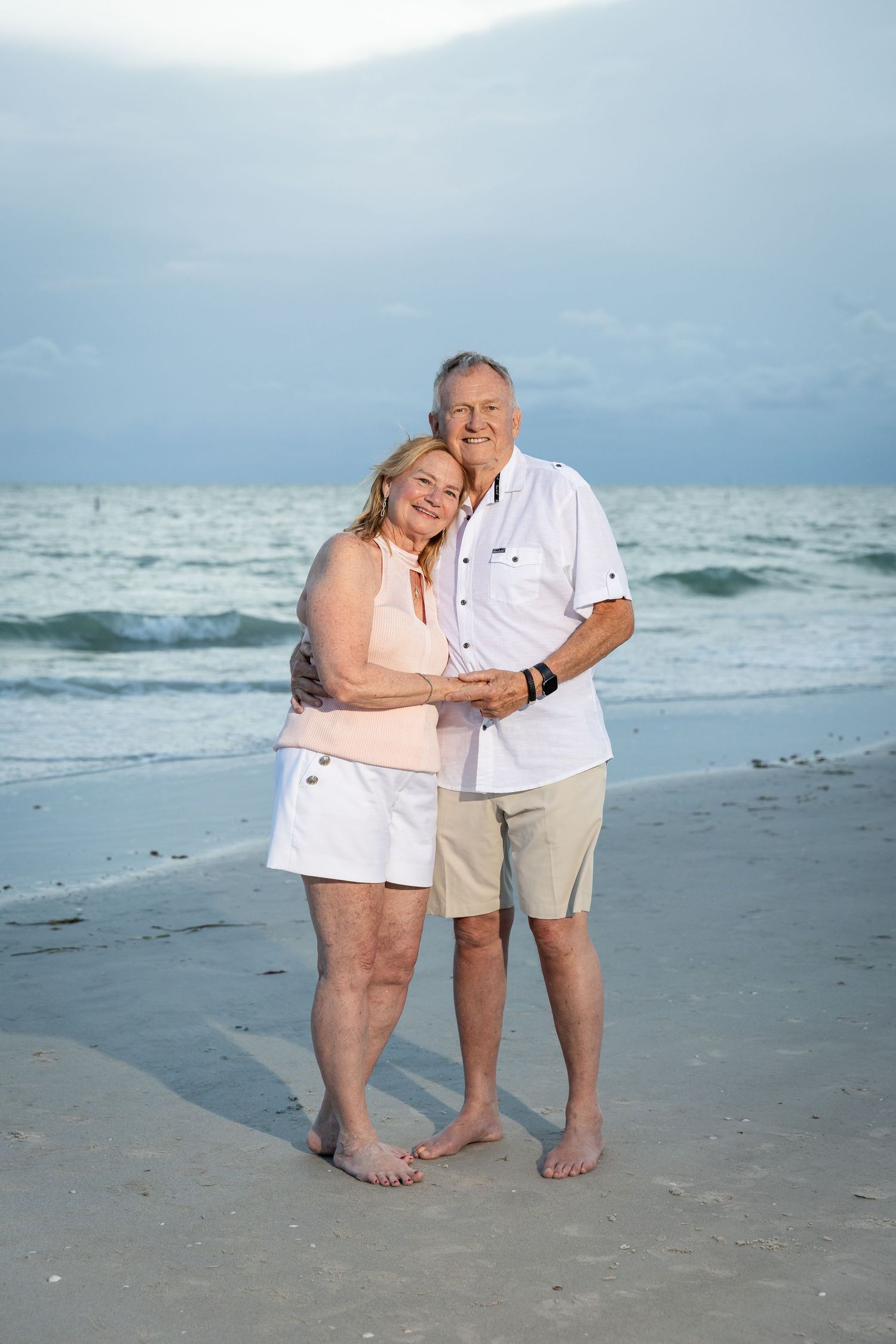 Happy senior couple embracing on a sandy beach at sunset, wearing casual summer clothes, with ocean waves in the background.