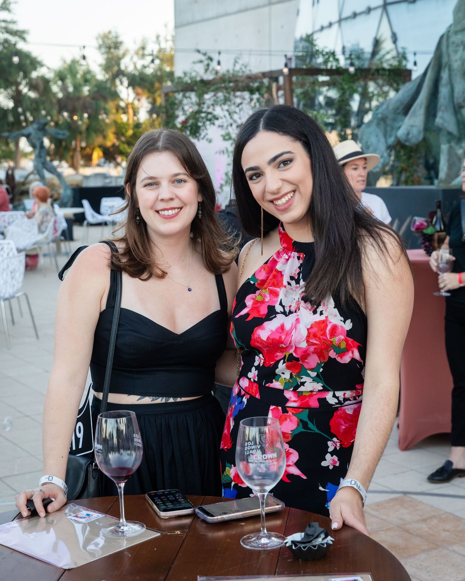 Two smiling women pose together at an outdoor evening event, holding glasses of red wine.