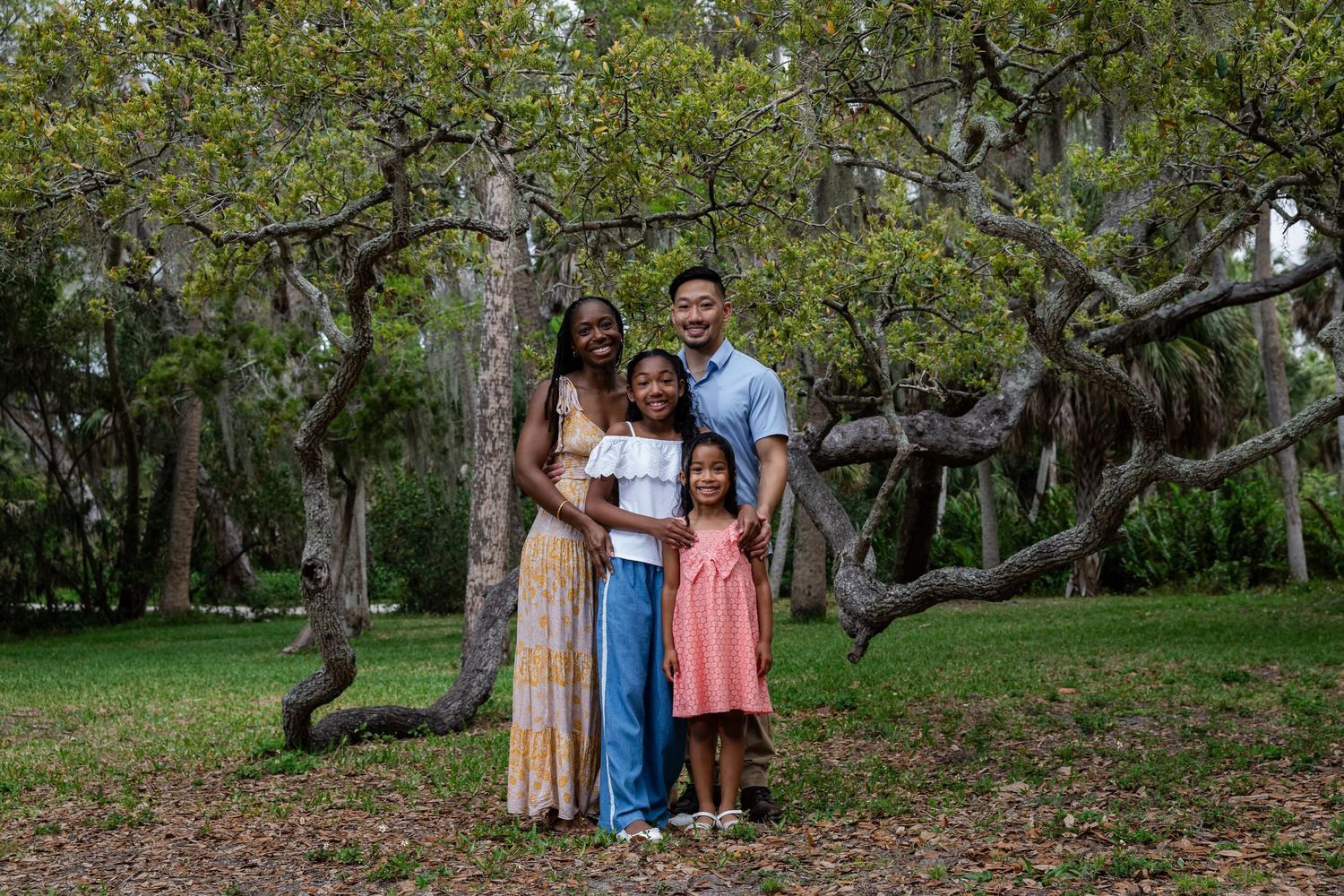 Happy family of four posing together under twisted trees in a lush green park setting.