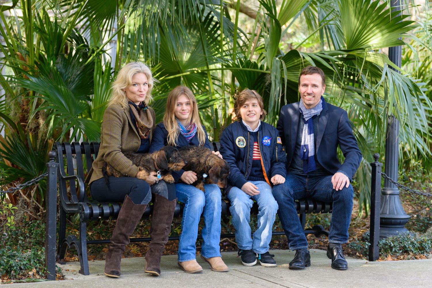 Family of four posing on a park bench surrounded by lush tropical palm trees outdoors.