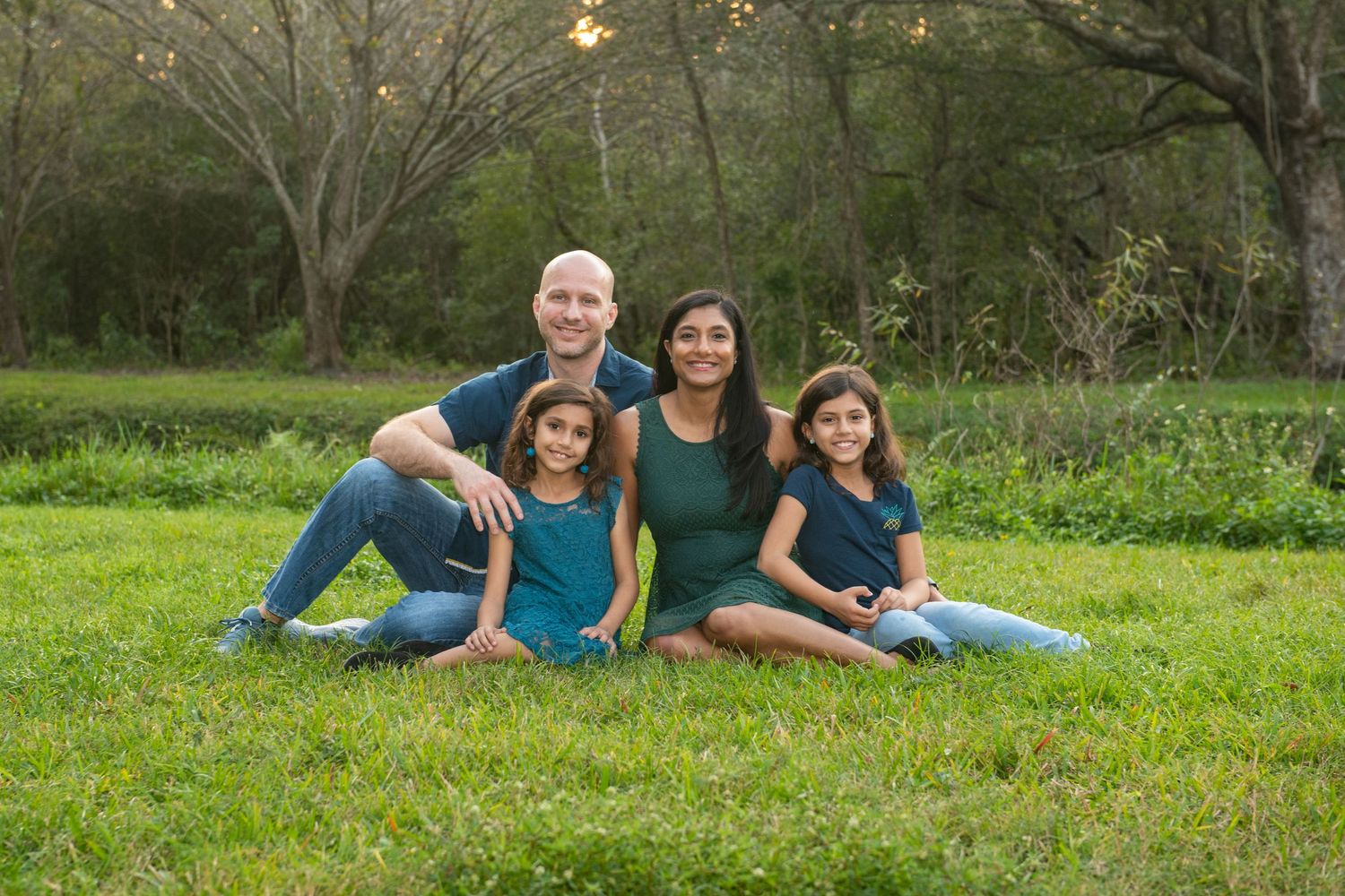 Family of five smiling outdoors on green grass surrounded by trees, wearing coordinated teal and blue outfits.