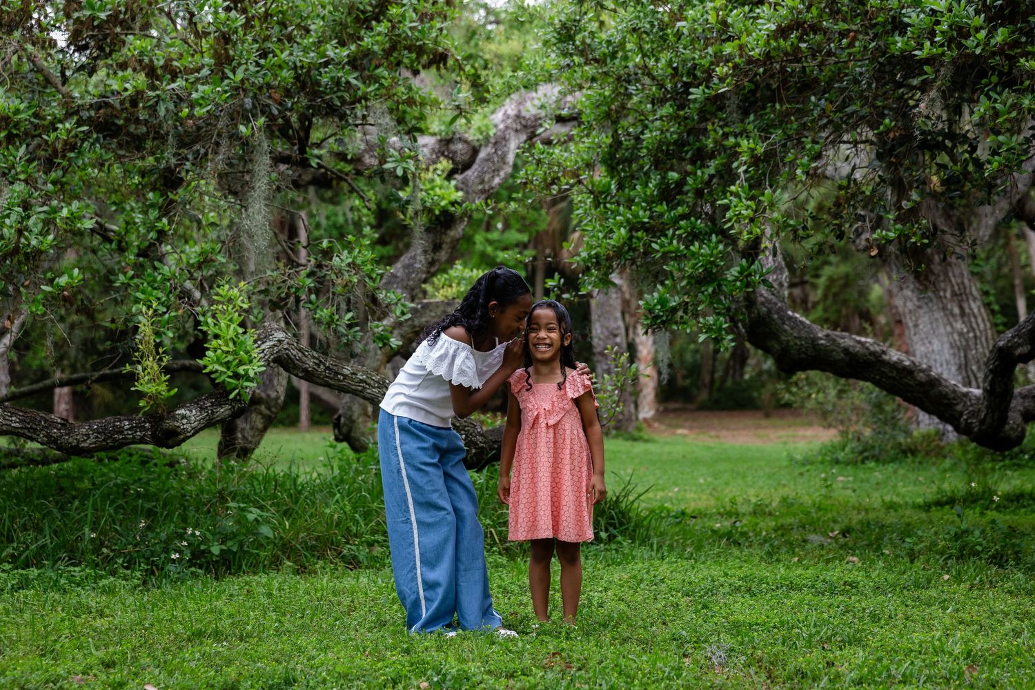 A woman leans down to kiss a young girl in a pink dress, standing together in a lush green park with mossy trees.